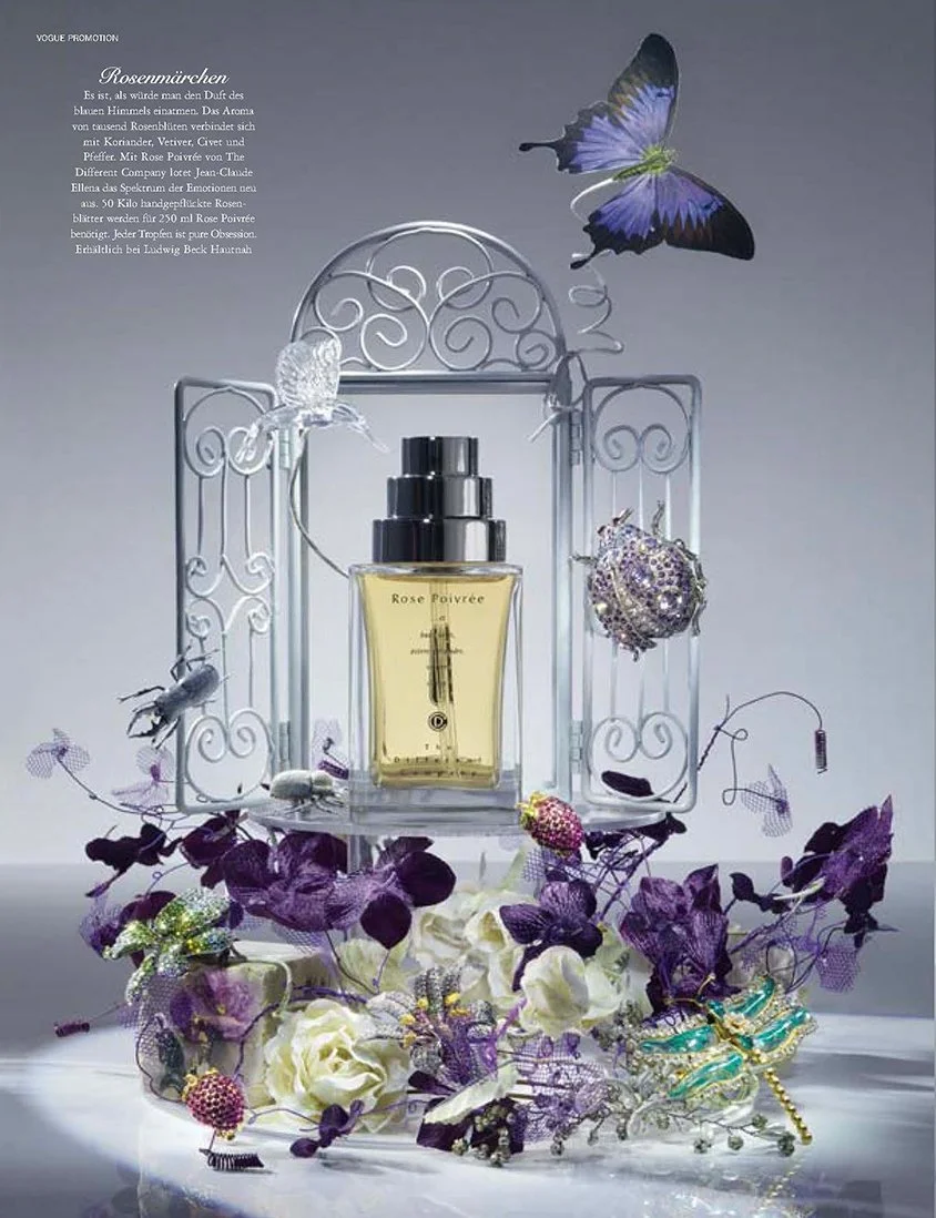 Stillleben-Fotografie von imagespy für VOGUE: Ein Parfum-Flakon von The Different Company in einem dekorativen silbernen Metall-Pavillon, umrahmt von violetten Orchideen, weißen Rosen und juwelenbesetzten Schmetterlingen für Ludwig Beck Hautnah.