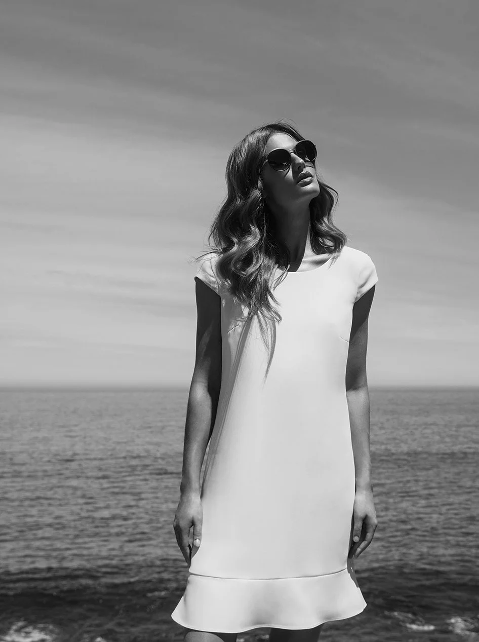 Schwarzweiß-Aufnahme eines Models in minimalistischem weißen Sommerkleid mit Sonnenbrille, stehend im Meer – People-Motiv aus comma SS2018, kreativ umgesetzt von imagespy.