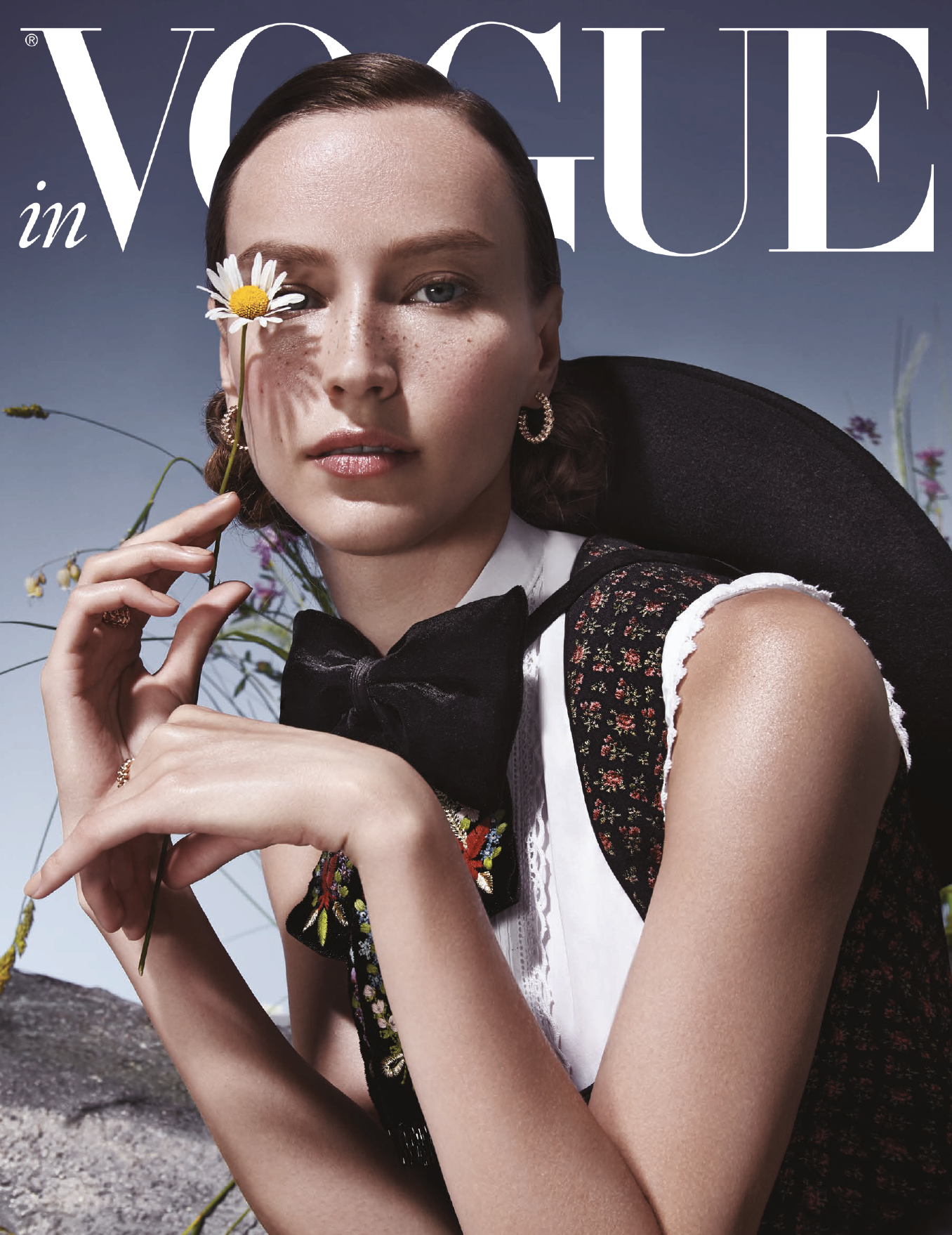 Model in traditioneller Tracht hält eine Margerite vors Gesicht, sitzt auf einem Stein vor einer hohen Wiese – moderne Trachteninszenierung von imagespy für Vogue und Lodenfrey.