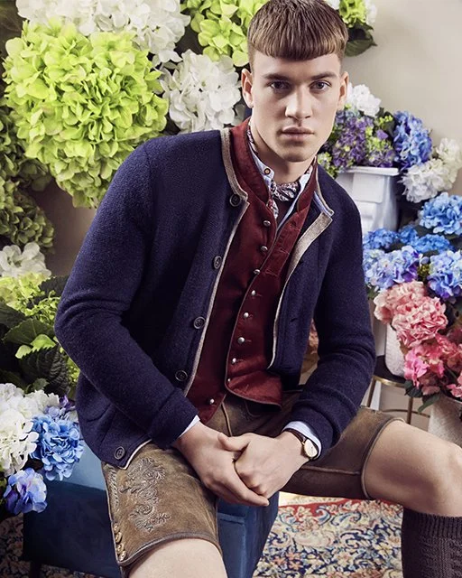 Male Model sitzt vor einer bunten Hortensienkulisse, trägt dunkelblaue Strickjacke und braune Lederhose. Kampagnenaufnahme von imagespy für die Trachtenmanufaktur München.