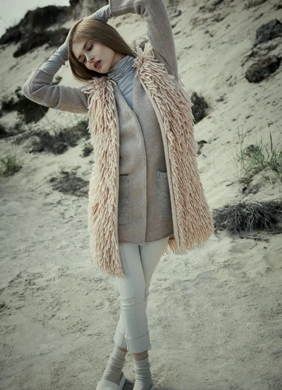 Model am Strand mit Boho-Fransenweste in Puder, greigem Schurwoll-Cardigan, cremefarbener 7/8-Hose und hellblauem Rollkragen-Shirt; umgesetzt von imagespy.