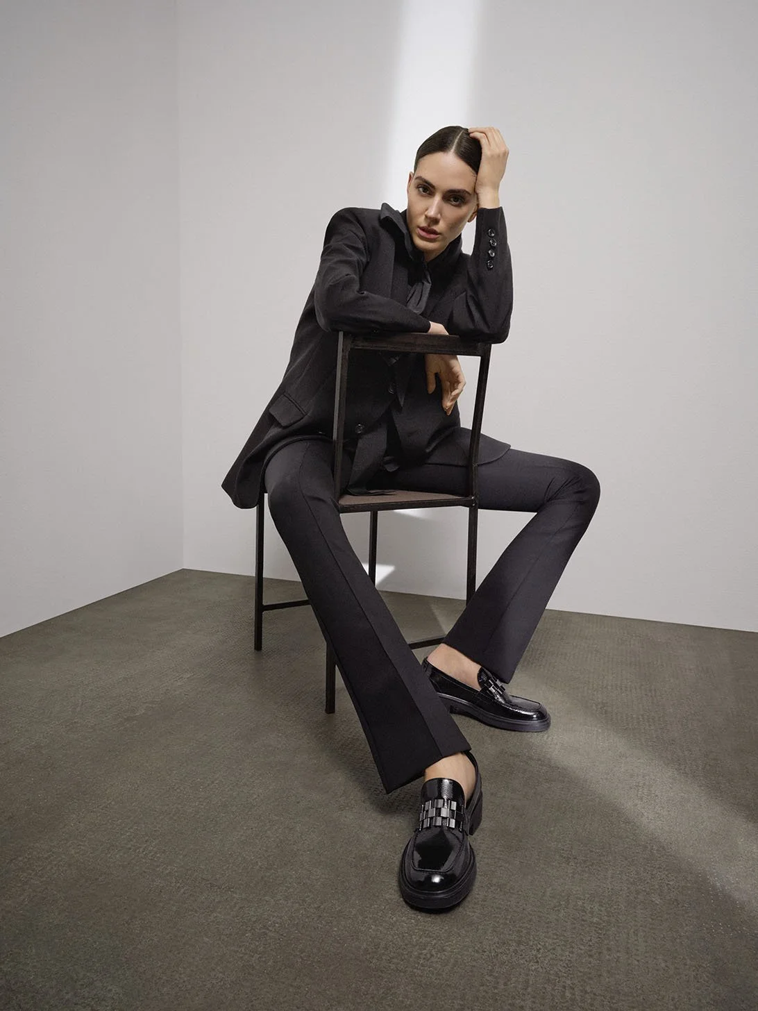 Högl Fall/Winter 2023: Model im Business-Outfit auf minimalistischem Stuhl mit schwarzen Högl Loafern, realisiert von imagespy für 360°-Kampagne.