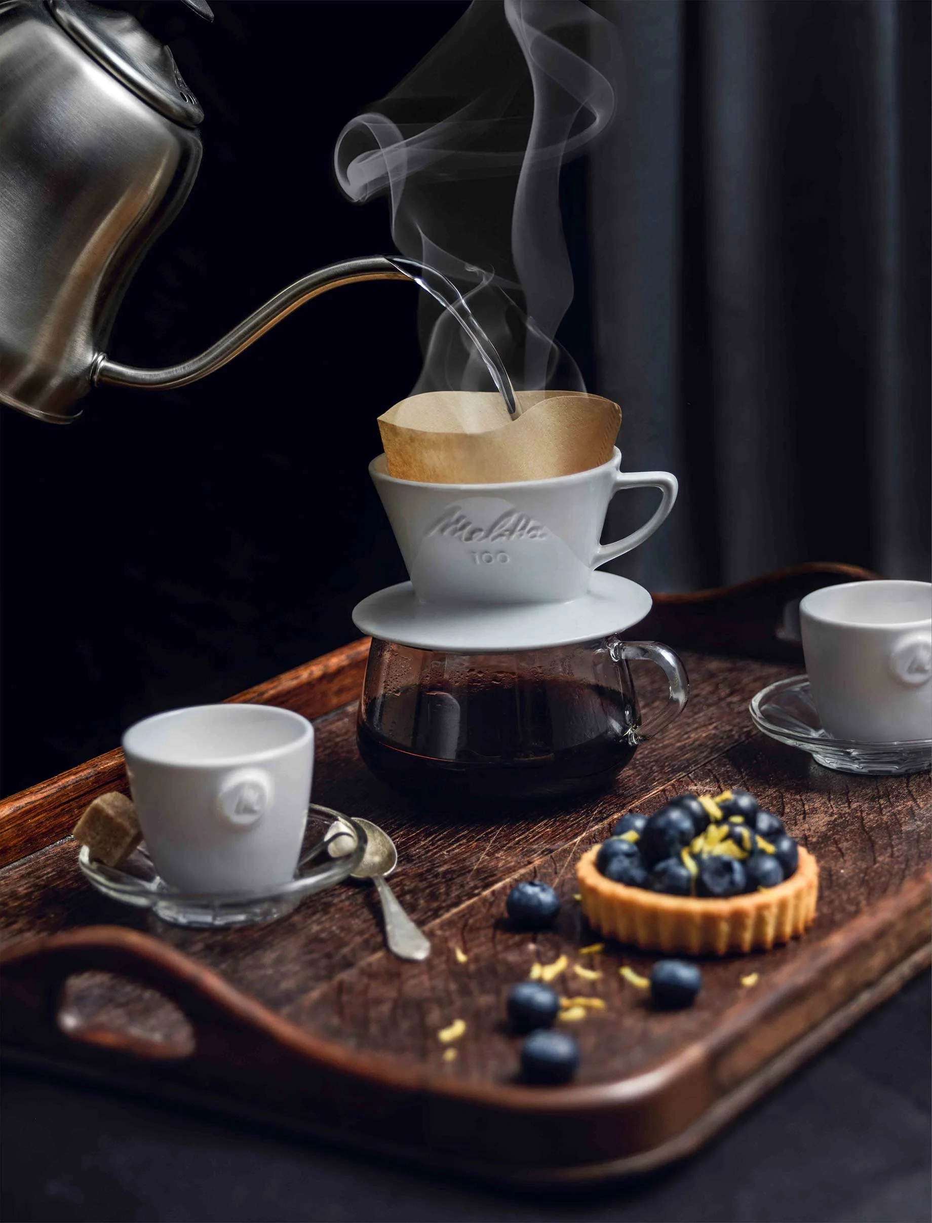 Klassischer Melitta Porzellan-Handfilter beim Aufbrühen auf einem rustikalen Holztablett, serviert mit einem Blaubeer-Törtchen – inszeniert von imagespy zum 111-jährigen Jubiläum.