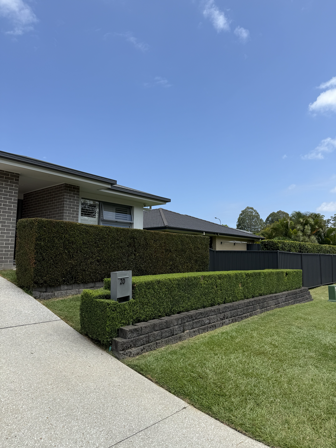 Front yard of a modern house with well-manicured hedge, grass, and a mailbox with the number 20, under a cloudy blue sky.