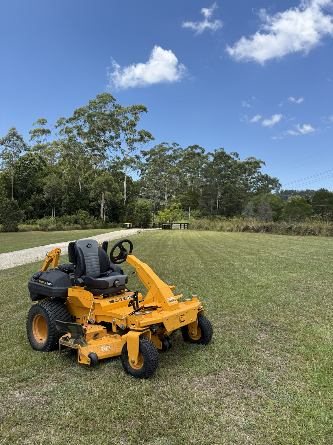 A yellow lawn mower on a well-maintained grass field, with a gravel path in the background and tall trees under a partly cloudy sky.