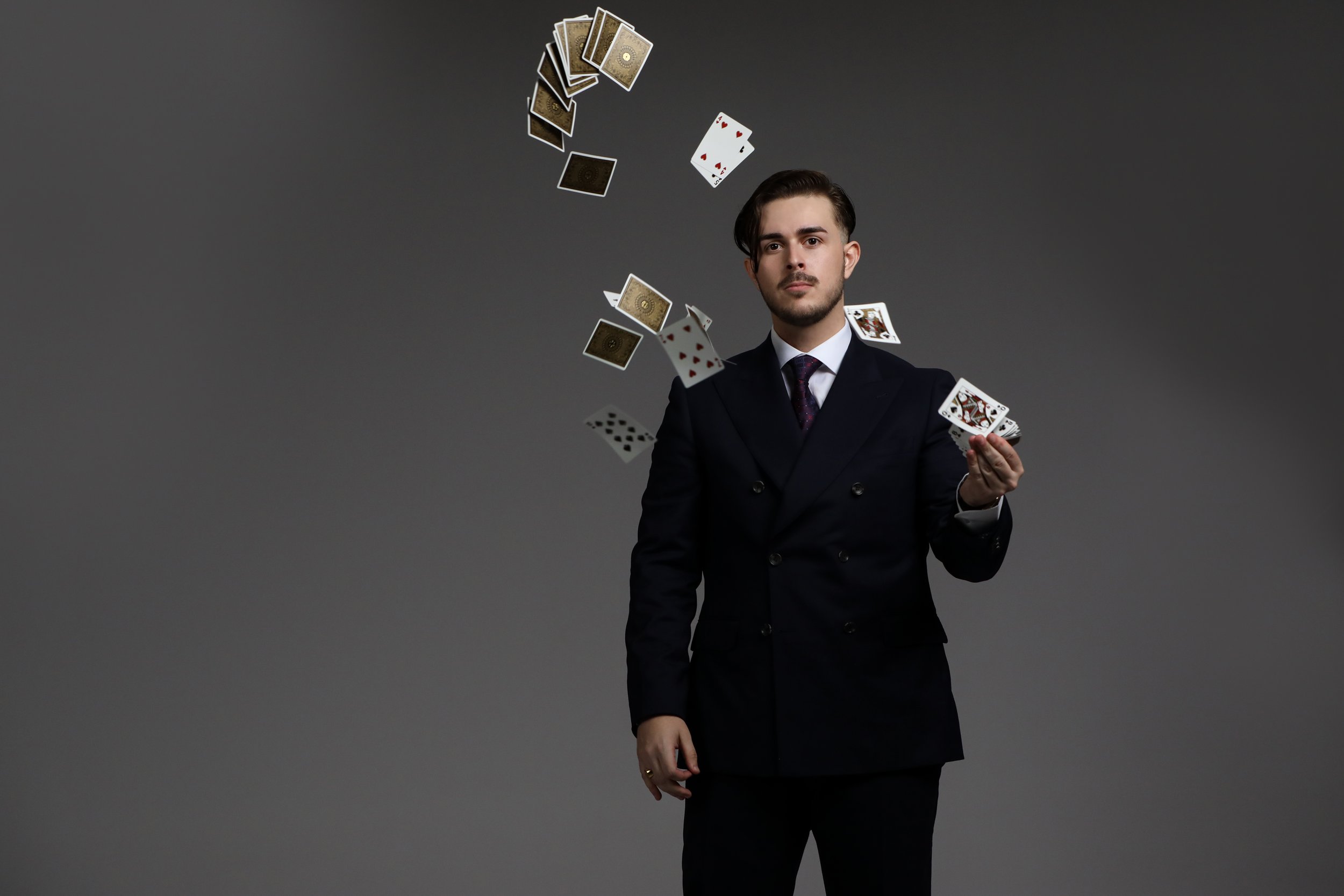 A man in a black suit holding a playing card with playing cards and a deck of cards flying around him against a gray background.