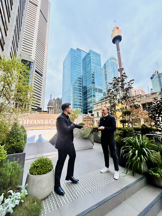 Two men in suits talking on a rooftop terrace in downtown Sydney, with city skyscrapers and the Sydney Tower Eye in the background, and potted plants around.