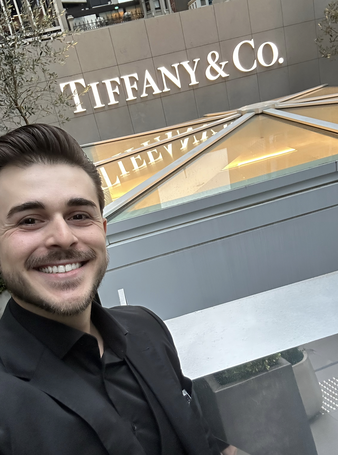 A smiling man taking a selfie in front of the Tiffany & Co. store sign on a city street.