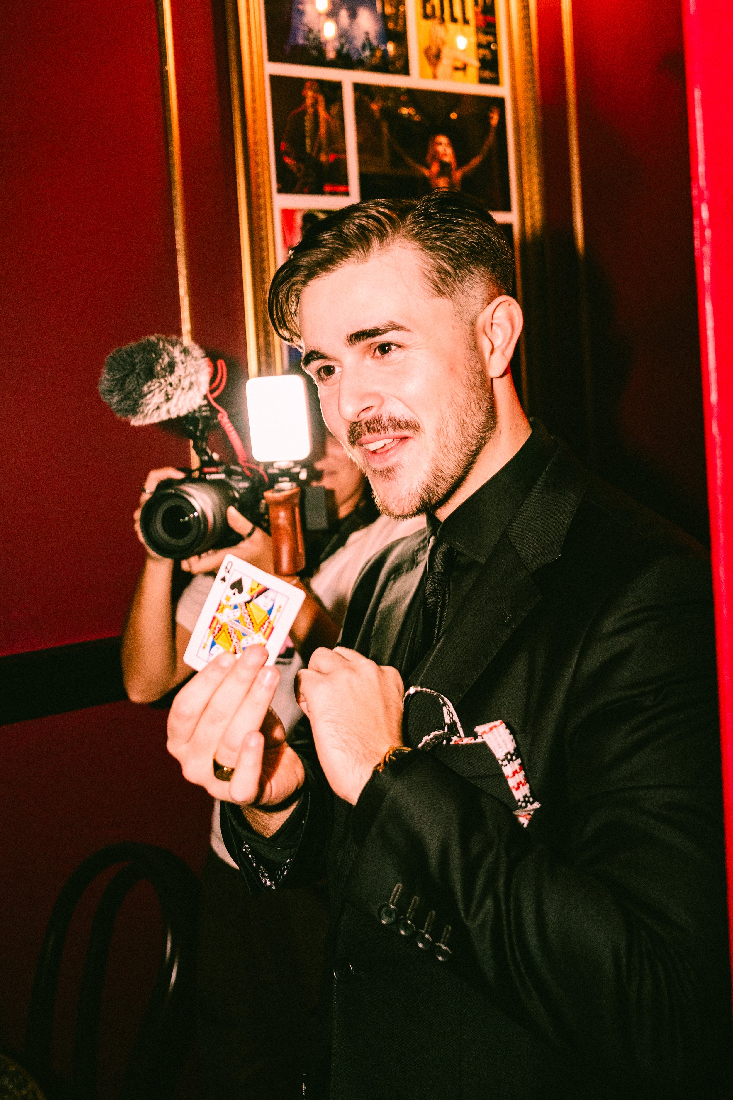 A man in a black suit holding a playing card, the Queen of Spades, at an indoor event with red and gold decor. In the background, a person is filming with a camera, and there are framed photographs or posters on the wall.