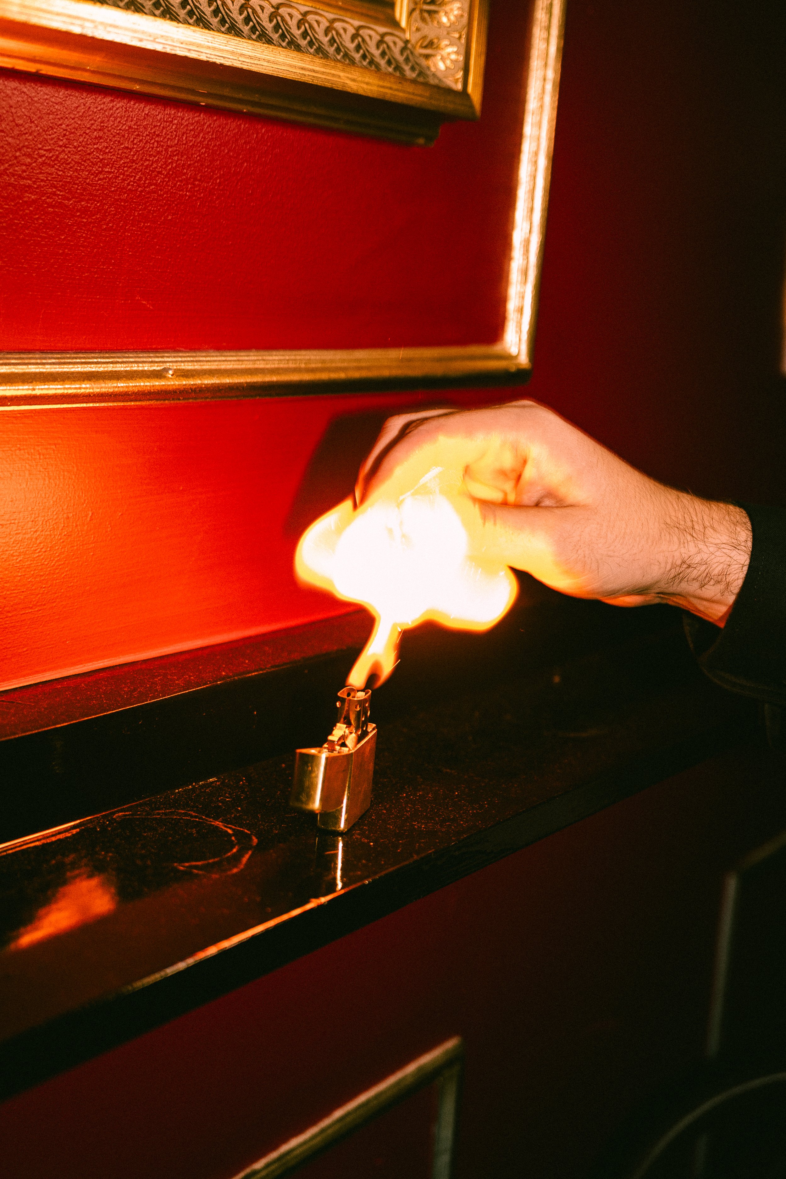 A person's hand holding a lighter near a flame in a dimly lit room with red walls and decorative gold trim.