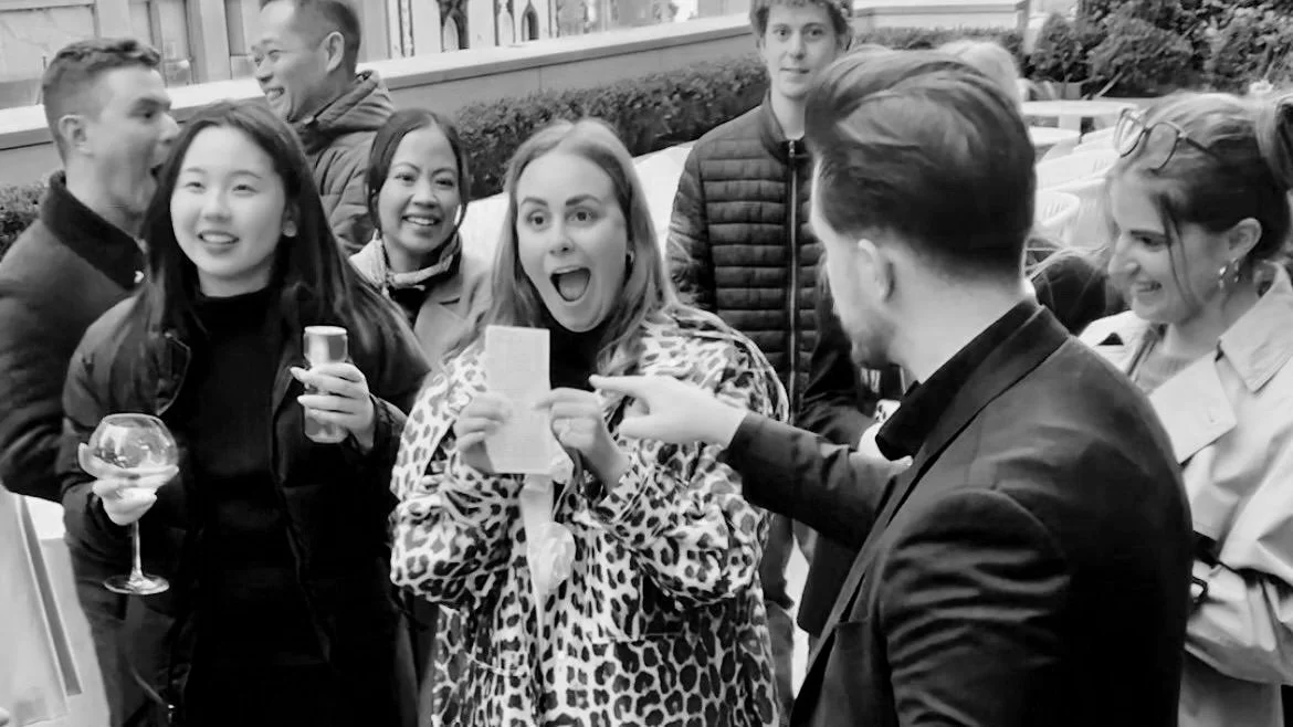 Group of people gathered outdoors, two women in foreground holding drinks and smiling, one woman showing a receipt or paper, a man pointing at it, others smiling and talking in the background.