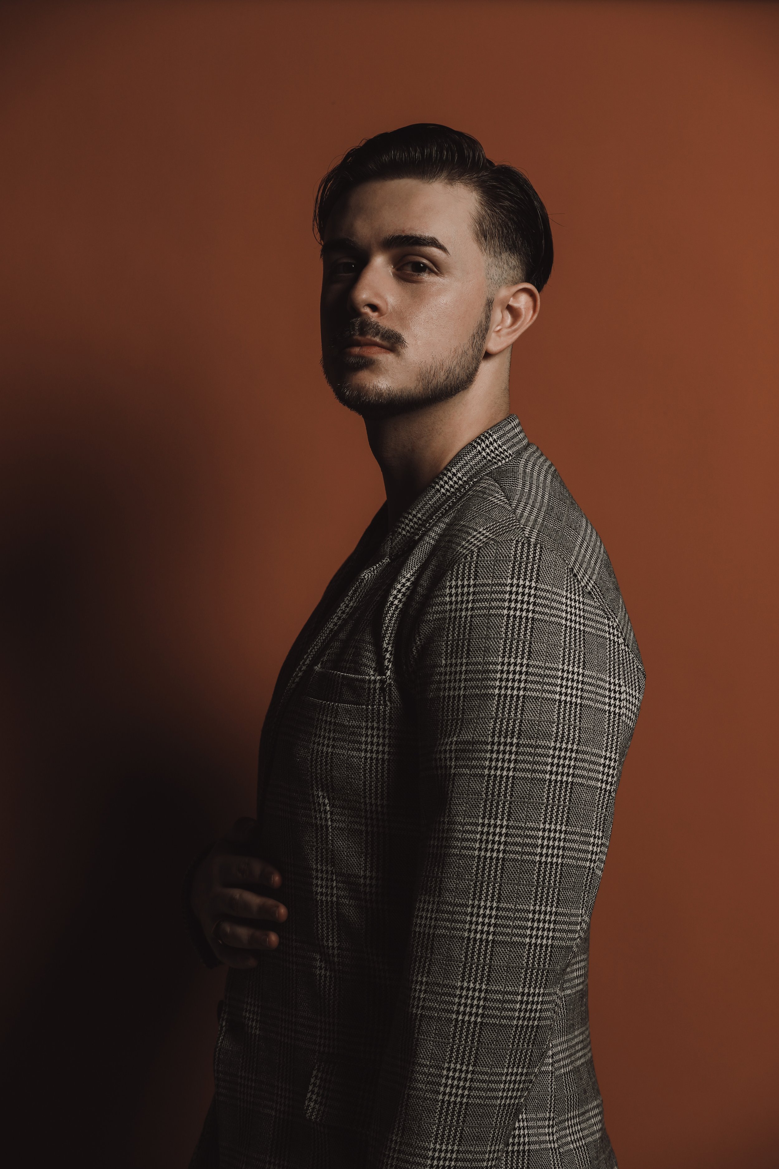Portrait of a man with styled dark hair, beard, wearing a checked blazer, standing against a gradient brown and orange background.