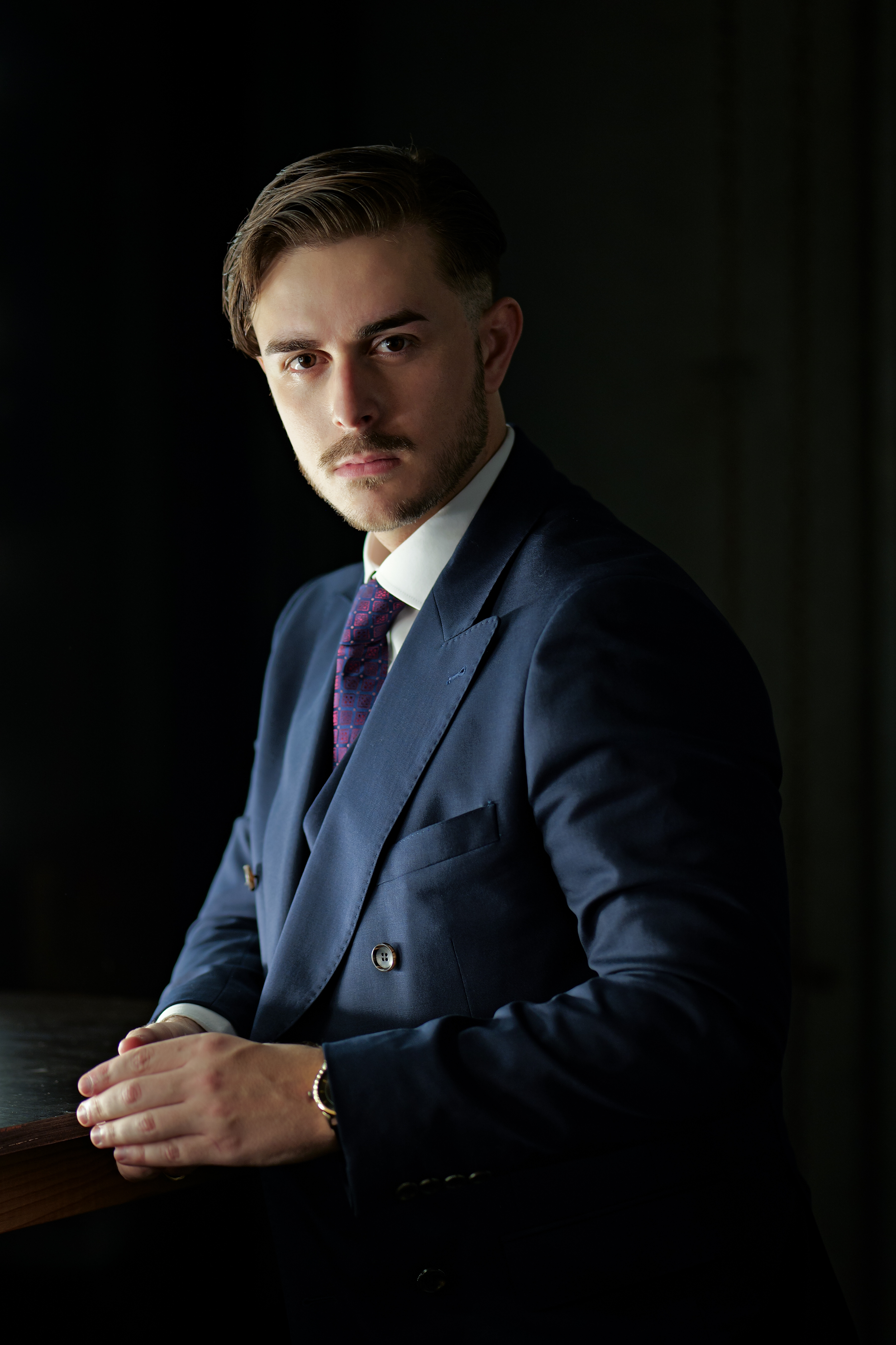A young man in a navy blue suit with a white shirt and purple tie, sitting at a wooden table, looking directly at the camera with a serious expression, in a dimly lit setting.