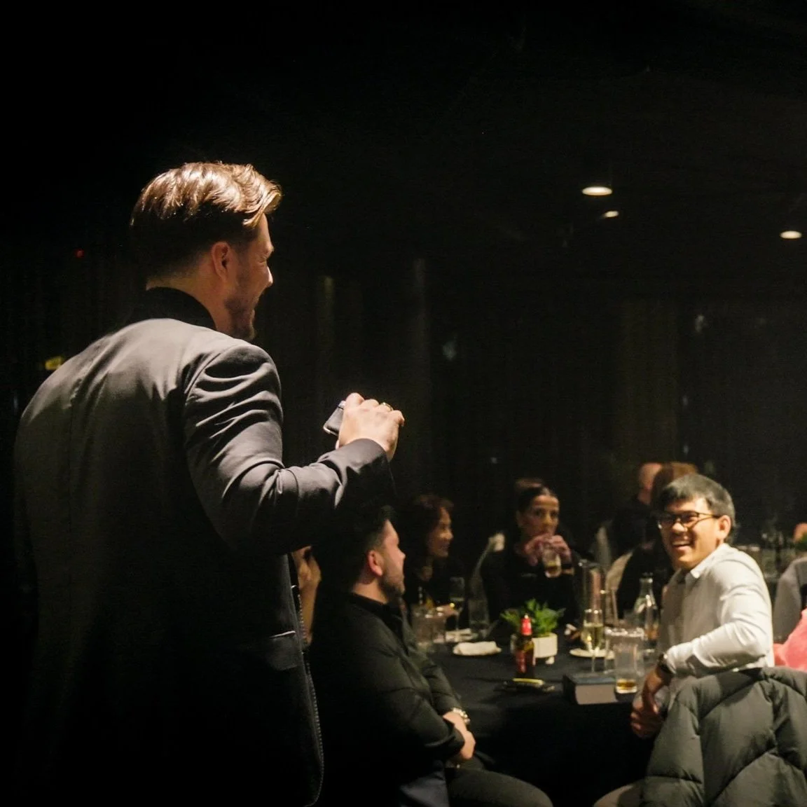 Man in a black jacket stands and talks to seated people at a social gathering or dinner, smiling and holding a drink.