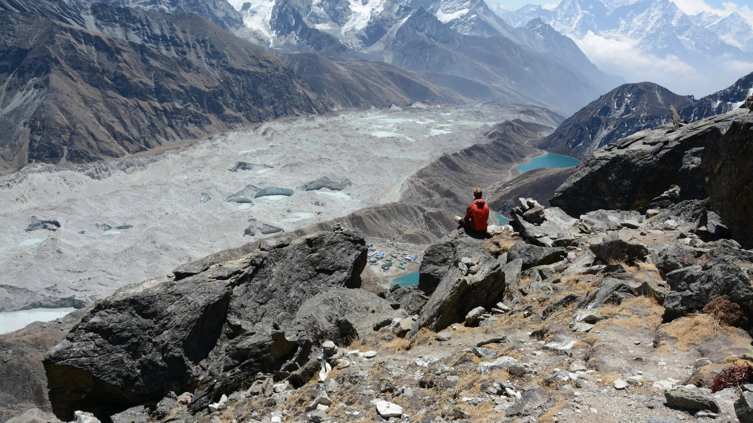 Mr Edward Saxby sitting on rocks overlooking a glacier valley with mountains in the distance, some with snow on their peaks.