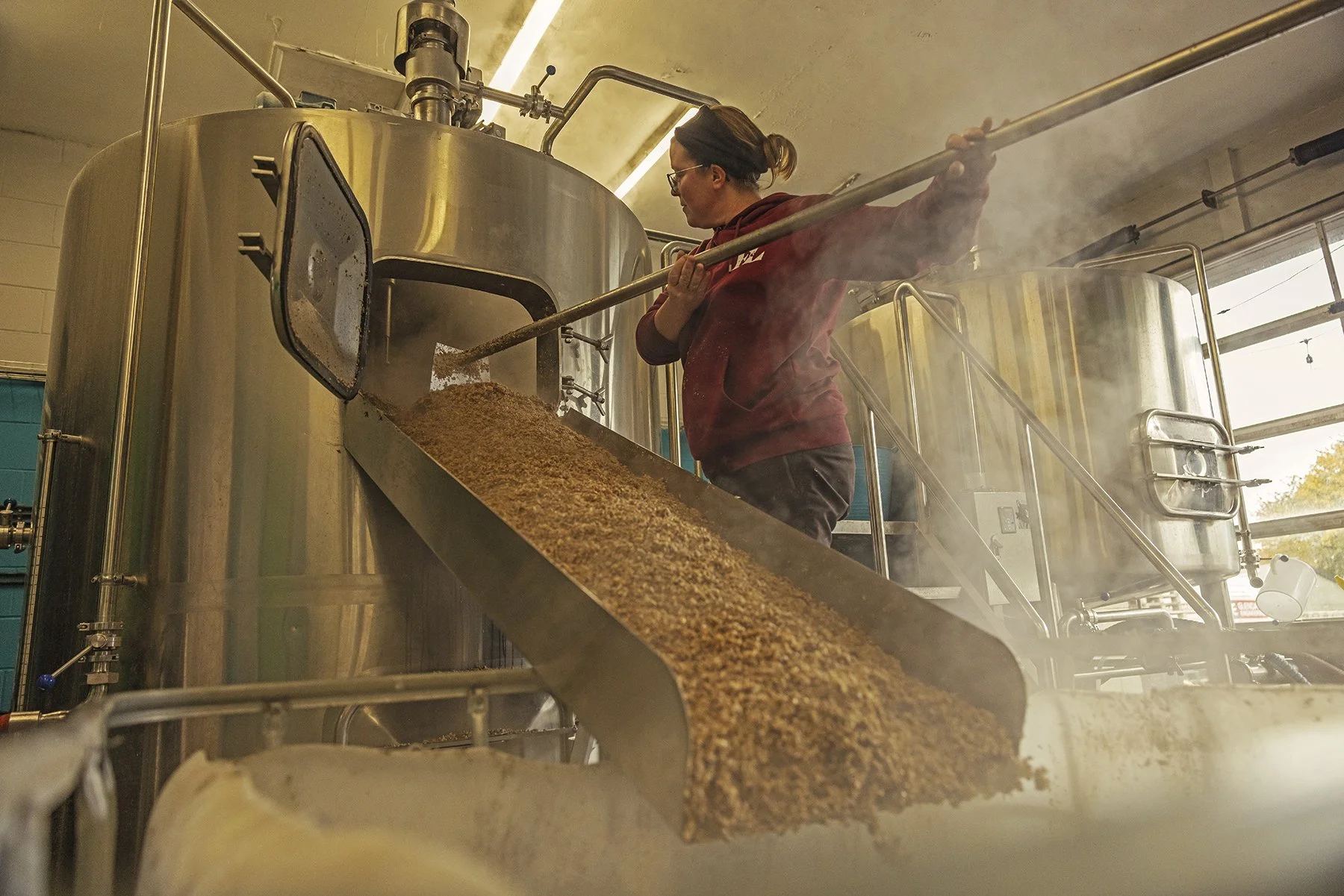 A woman in a maroon hoodie and glasses is working with a large stainless steel brewing tank, pouring a grain mixture onto a conveyor belt in a brewing or food processing facility.