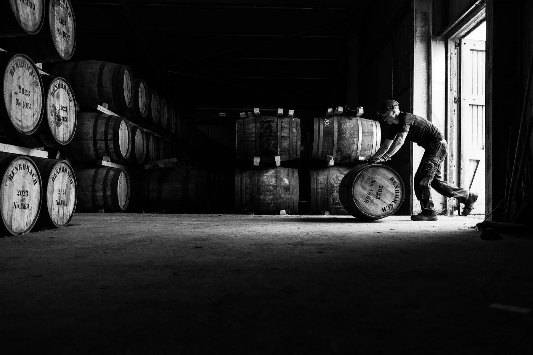 A person rolling a wooden barrel inside a dimly lit warehouse, surrounded by stacked barrels on racks.