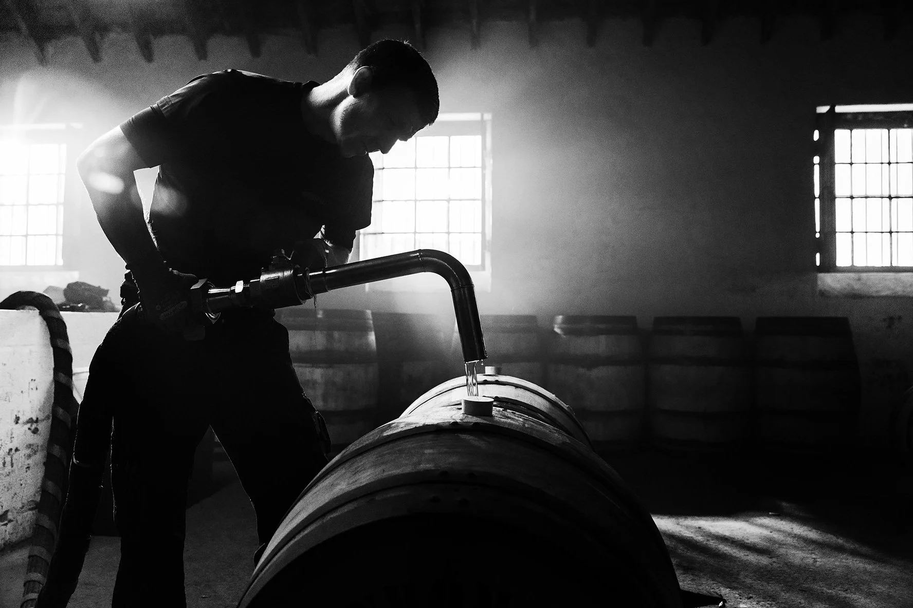 Silhouette of a person filling a wooden barrel with liquid through a metal pipe, in a dimly lit distillery or brewery with barrels in the background.