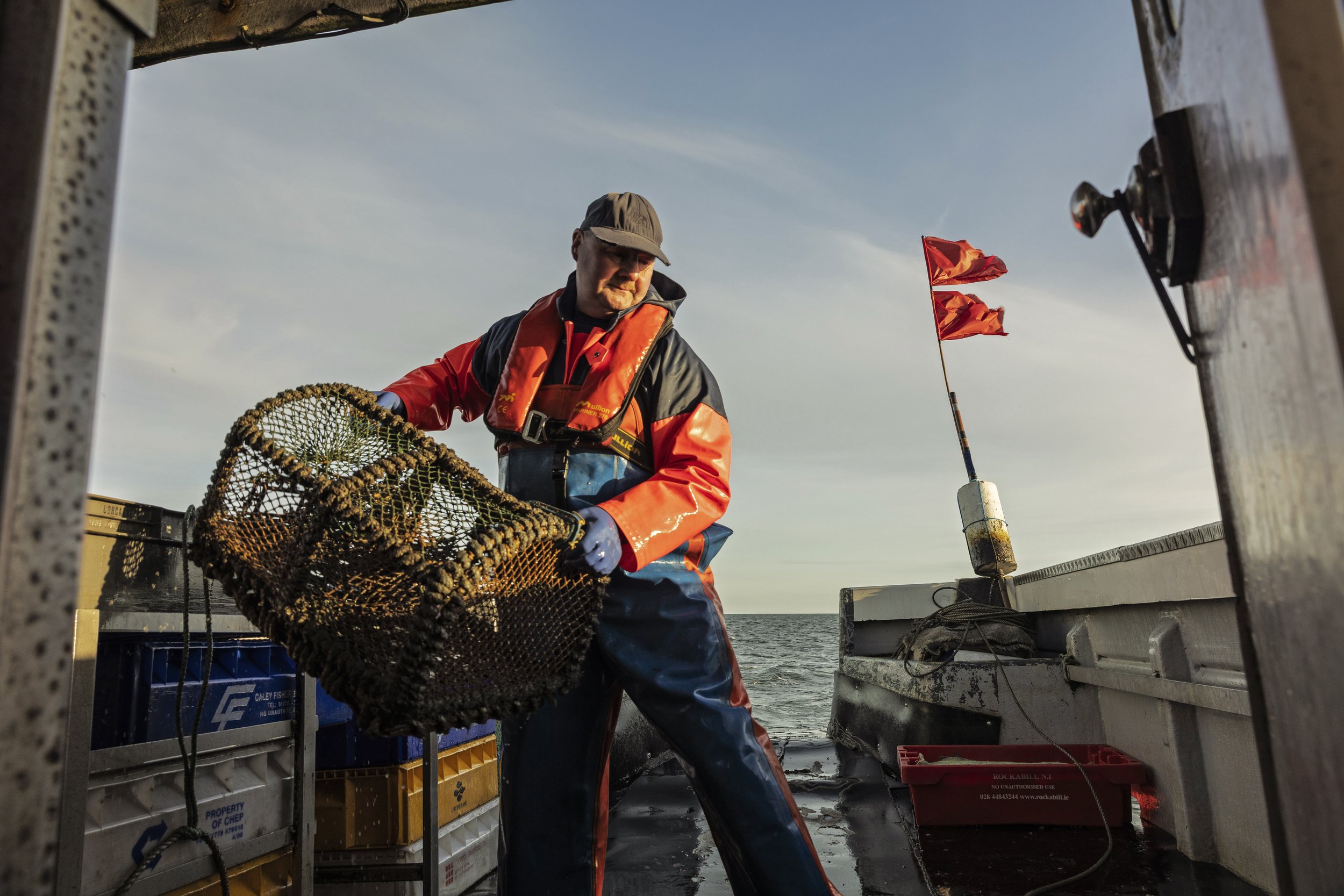 Man on a boat handling a lobster trap, wearing waterproof gear, with a red flag on a pole and ocean in the background.