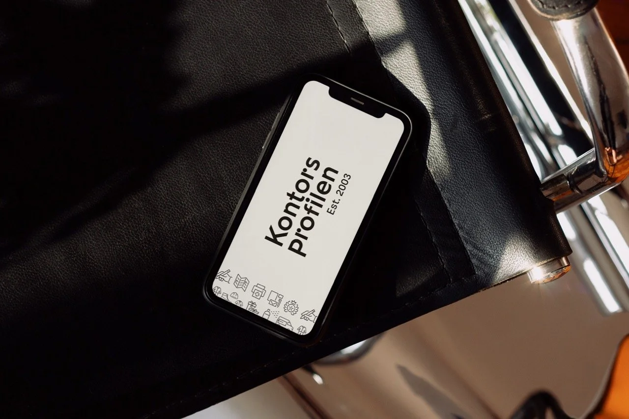 En smartphone ligger på en svart, skinnliknande yta som visar en hemsida för Kontoris Pffilien, med ikoner längst ner för olika funktioner.