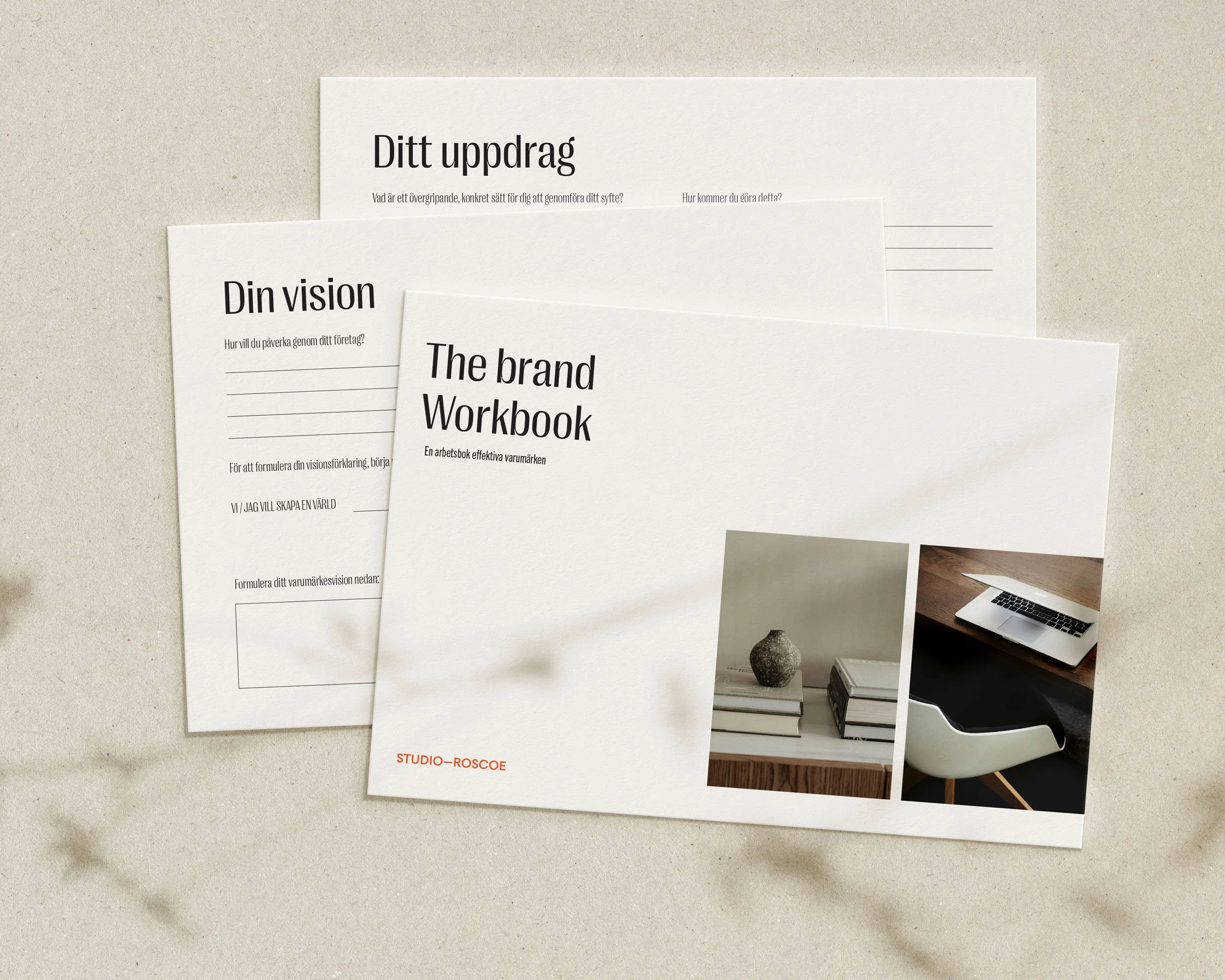 En arbetsbok med texterna "The brand Workbook", apart från ett blad med rubriken "Ditt uppdrag" och ett annat med "Din vision" samt ett foto av ett keramikvas och några böcker, ett datorfodral och en laptop på ett skrivbord.