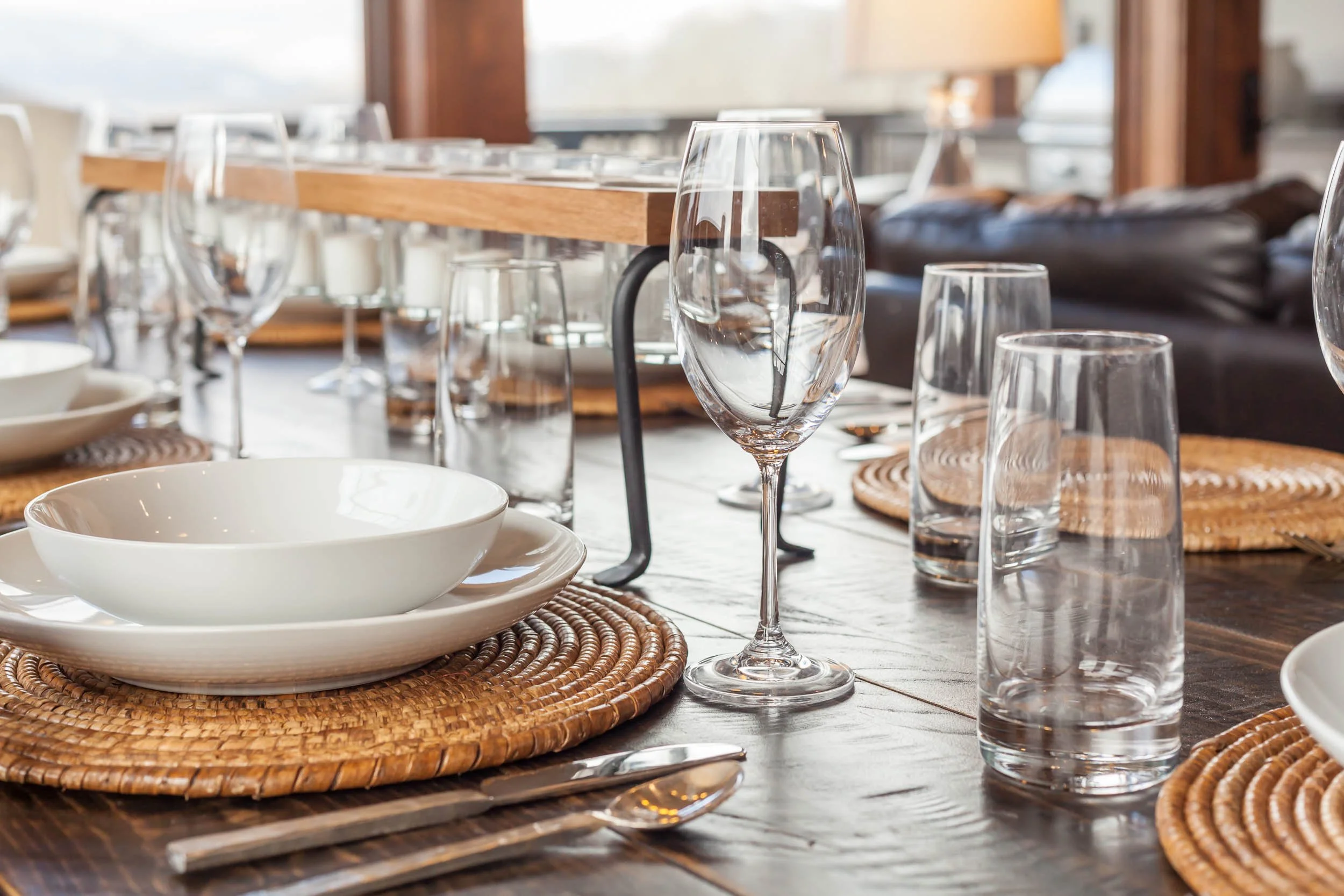 Empty dinner table set with white bowls, clear glasses, cutlery, woven placemats, and a wooden centerpiece in a well-lit room.