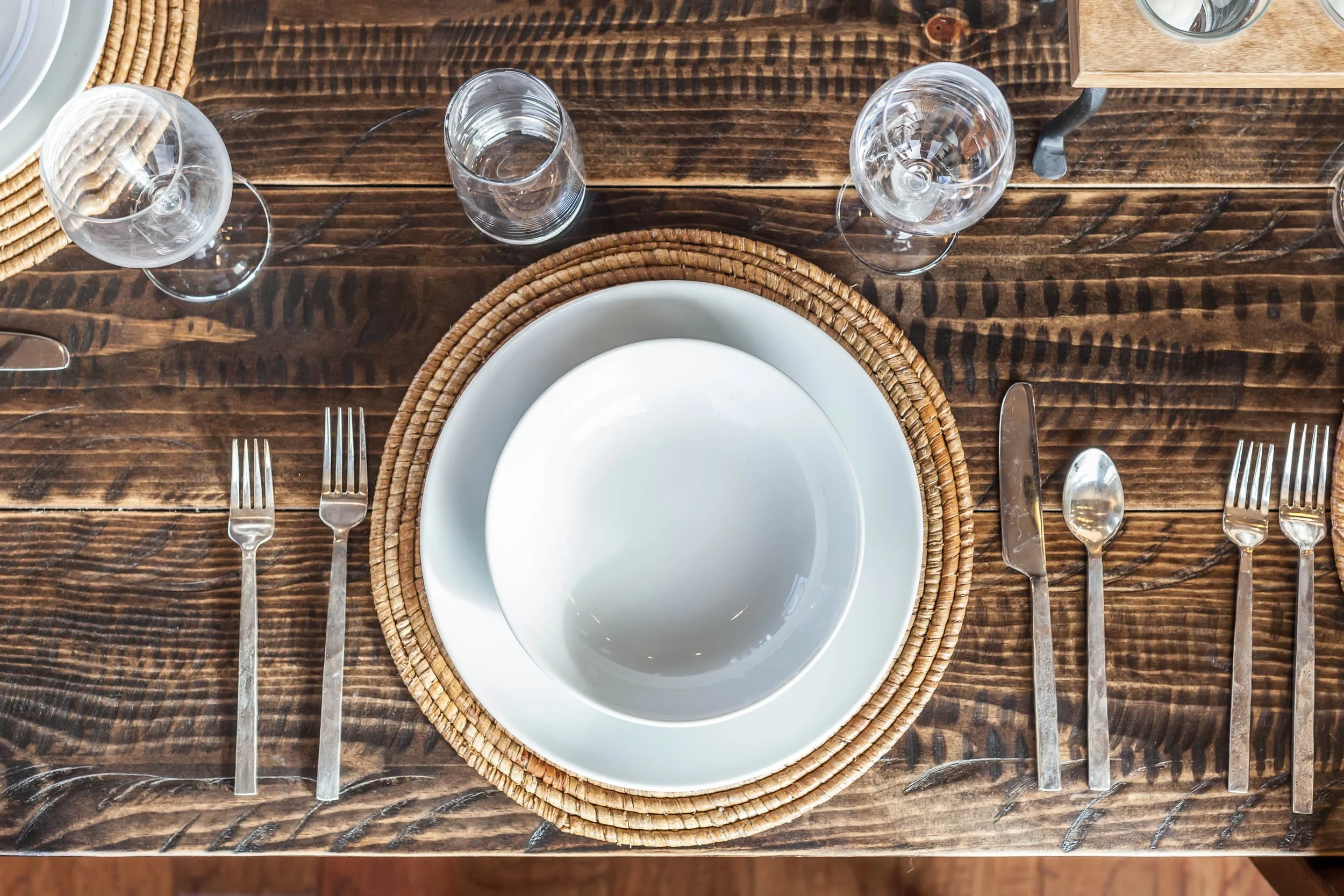 A top view of a table setting with a white bowl on a white plate, placed on a woven placemat, with silverware including two forks, a knife, and a spoon, alongside three wine glasses and two water glasses on a dark wooden table.