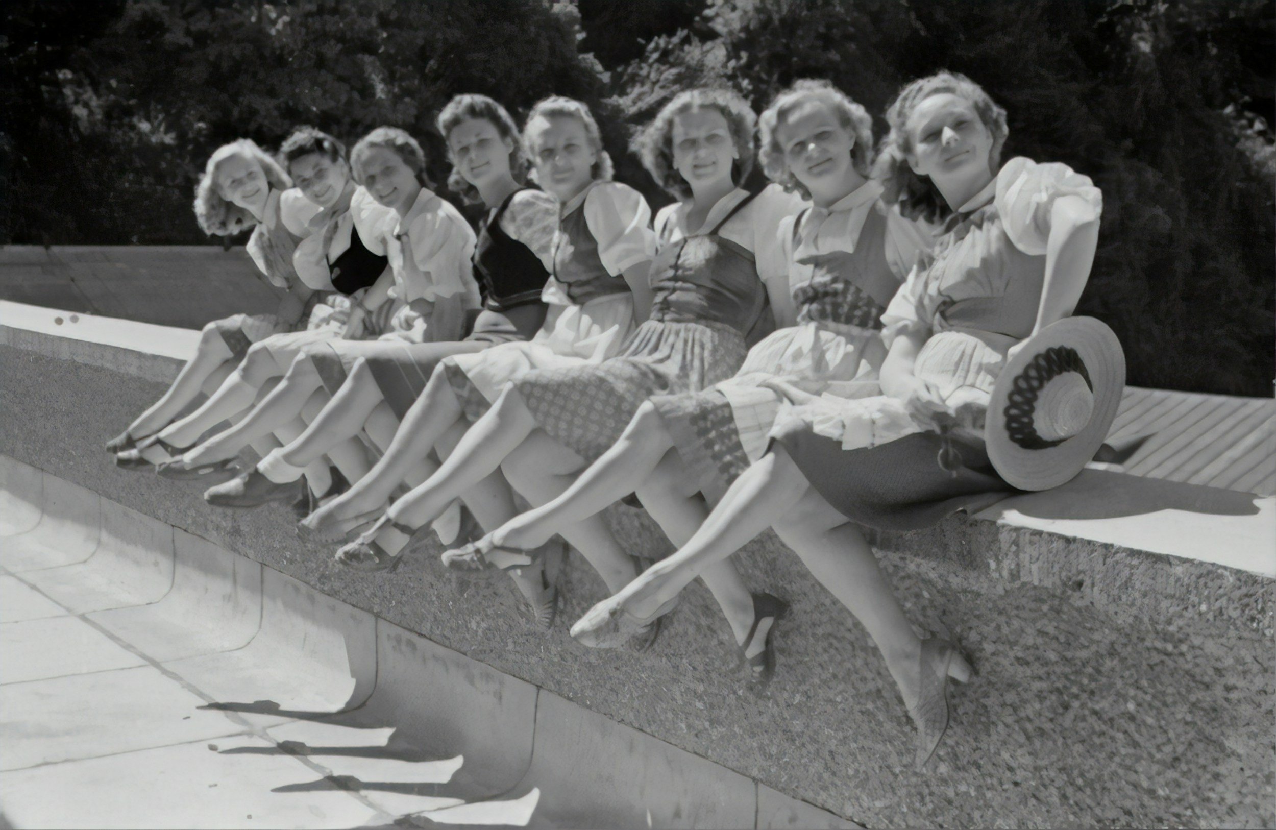 Black and white photo of eight women sitting on a park bench in vintage clothing, with trees in the background.