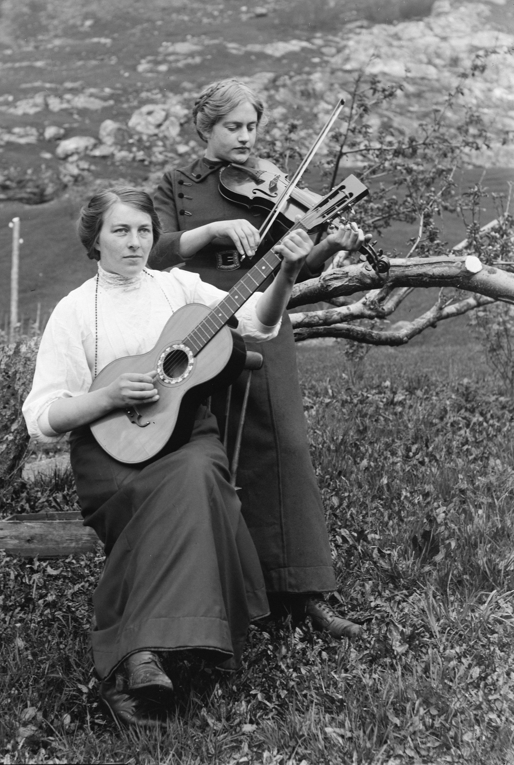 Two women outdoors playing string instruments, one with a guitar and the other with a violin, near a tree and rocks in the background.