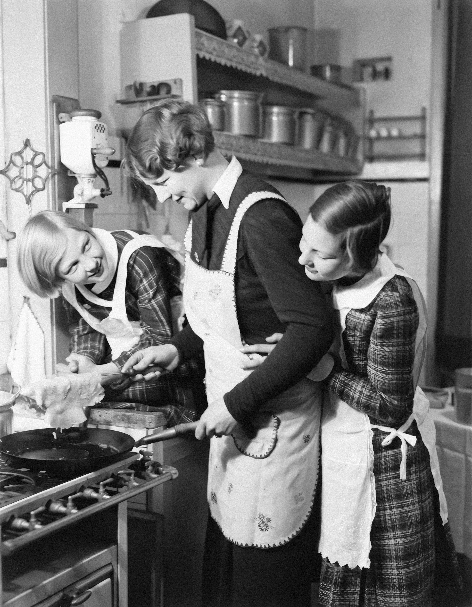 Three women wearing aprons cooking together in a kitchen, with pots and shelves in the background.