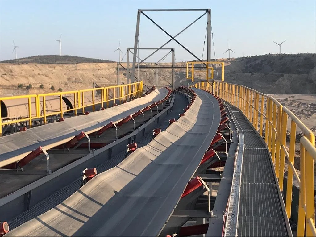 Industrial conveyor belt system with yellow safety railings, situated in a mining or quarry area, with wind turbines visible in the background.