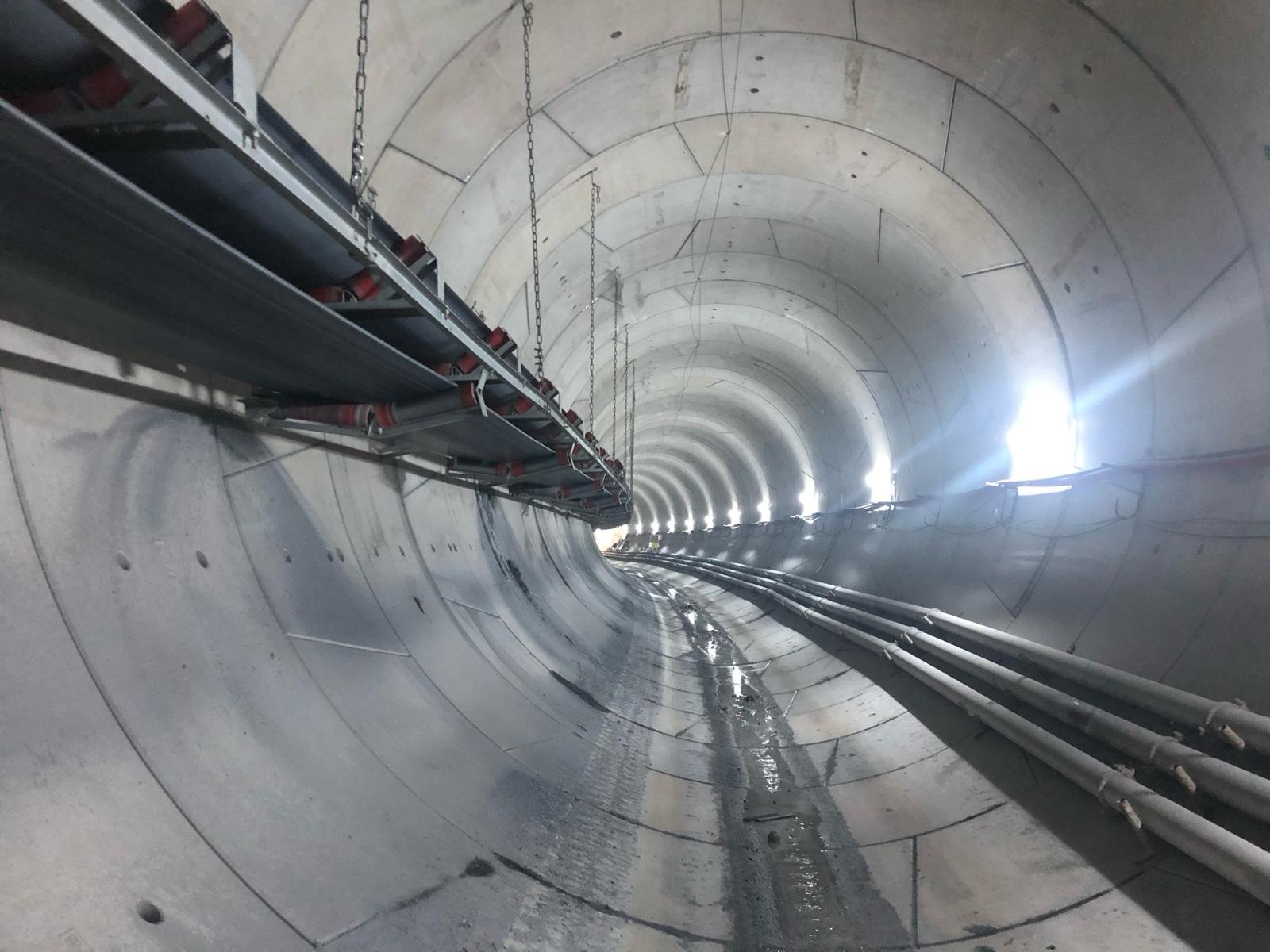 Interior of a concrete tunnel under construction with utility pipes and lights along the walls.