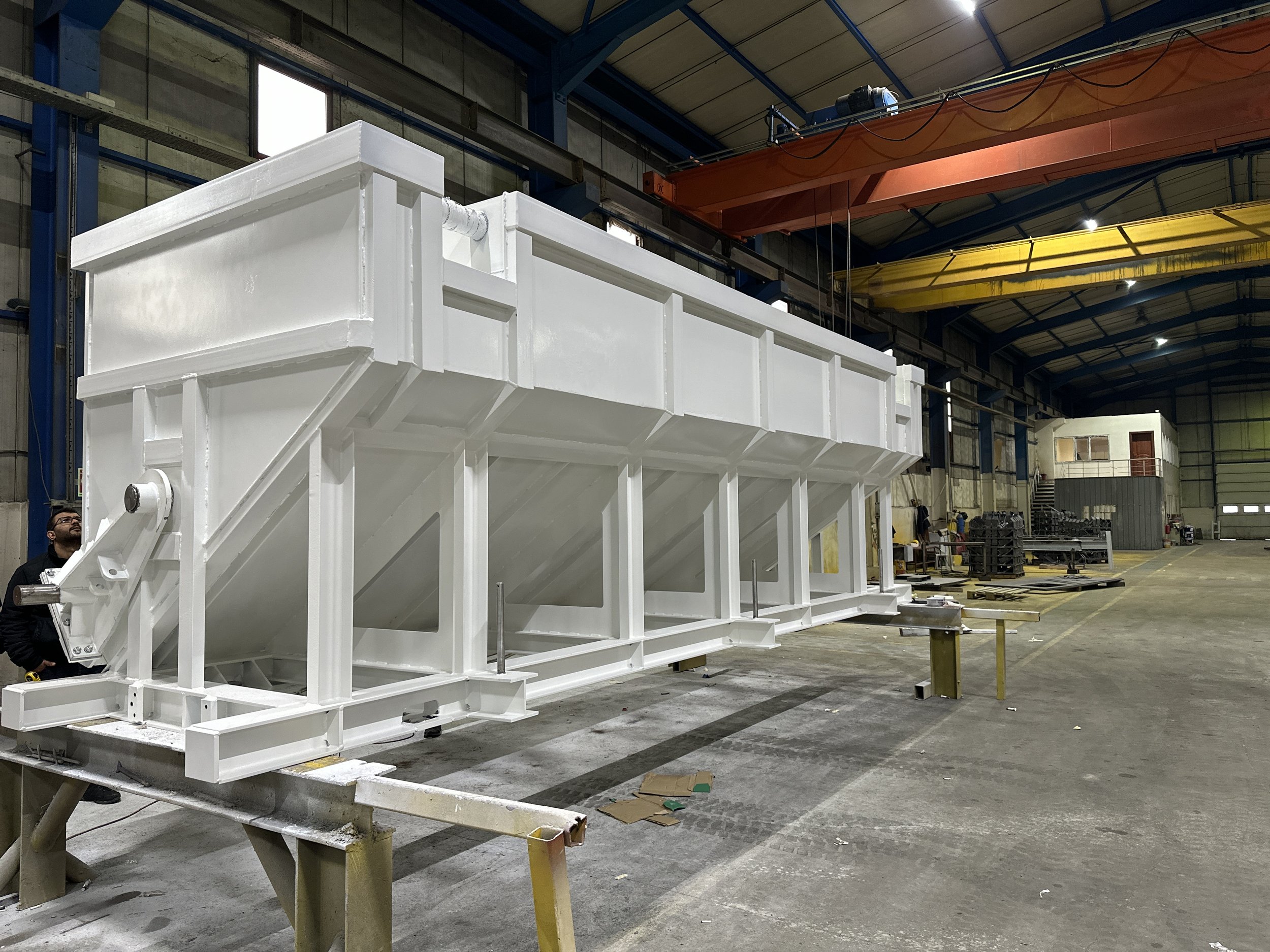 Large white industrial hopper inside a factory setting, supported by a metal frame, with a person nearby for scale.