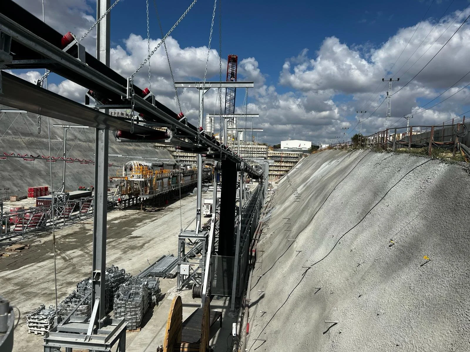 Construction site with heavy machinery, scaffolding, and equipment, surrounded by a sloped dirt embankment under a cloudy sky. Electrical cables are visible overhead.