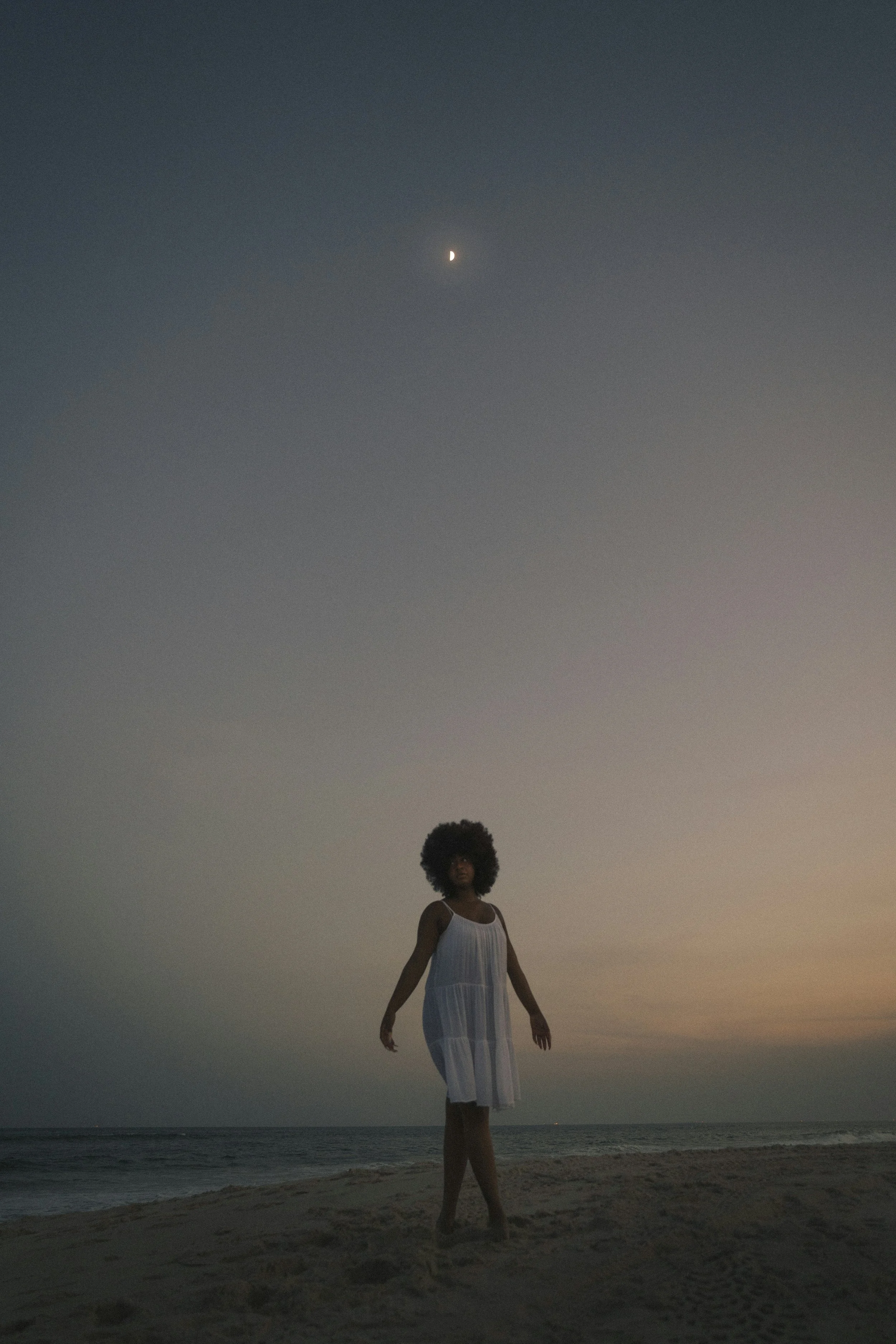 Femme africaine marchant sur la plage au coucher du soleil sous la lune, vêtue de blanc, symbole de féminité sacrée, de purification et de rituel lunaire.
