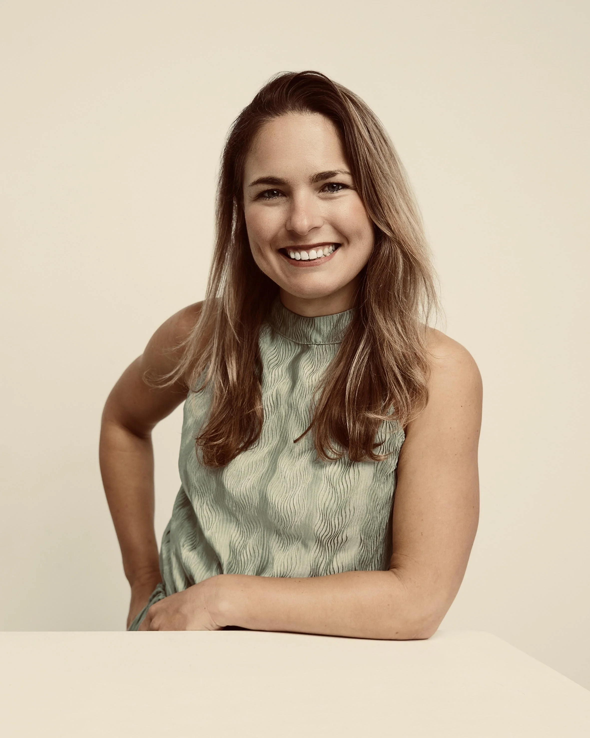 A woman with long, wavy brown hair, smiling, wearing a sleeveless green top, sitting with one arm on a white table against a plain beige background.