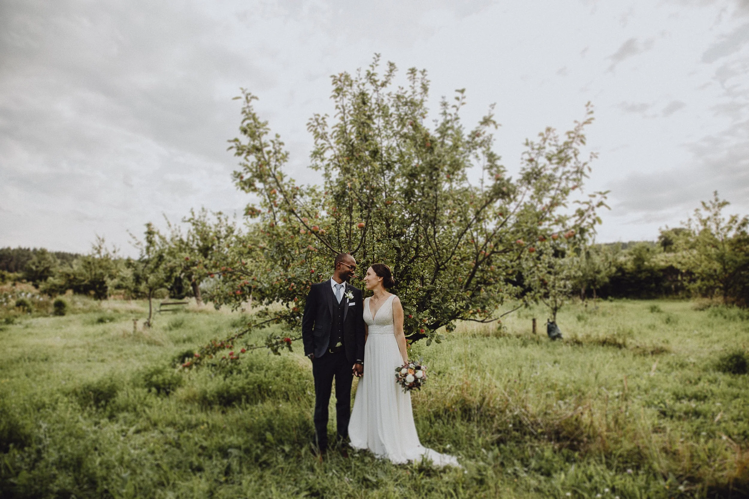 Ein glückliches Brautpaar steht in einem Apfelhain, Händchen haltend, die Braut hält einen Blumenstrauß, bewölkter Himmel im Hintergrund.