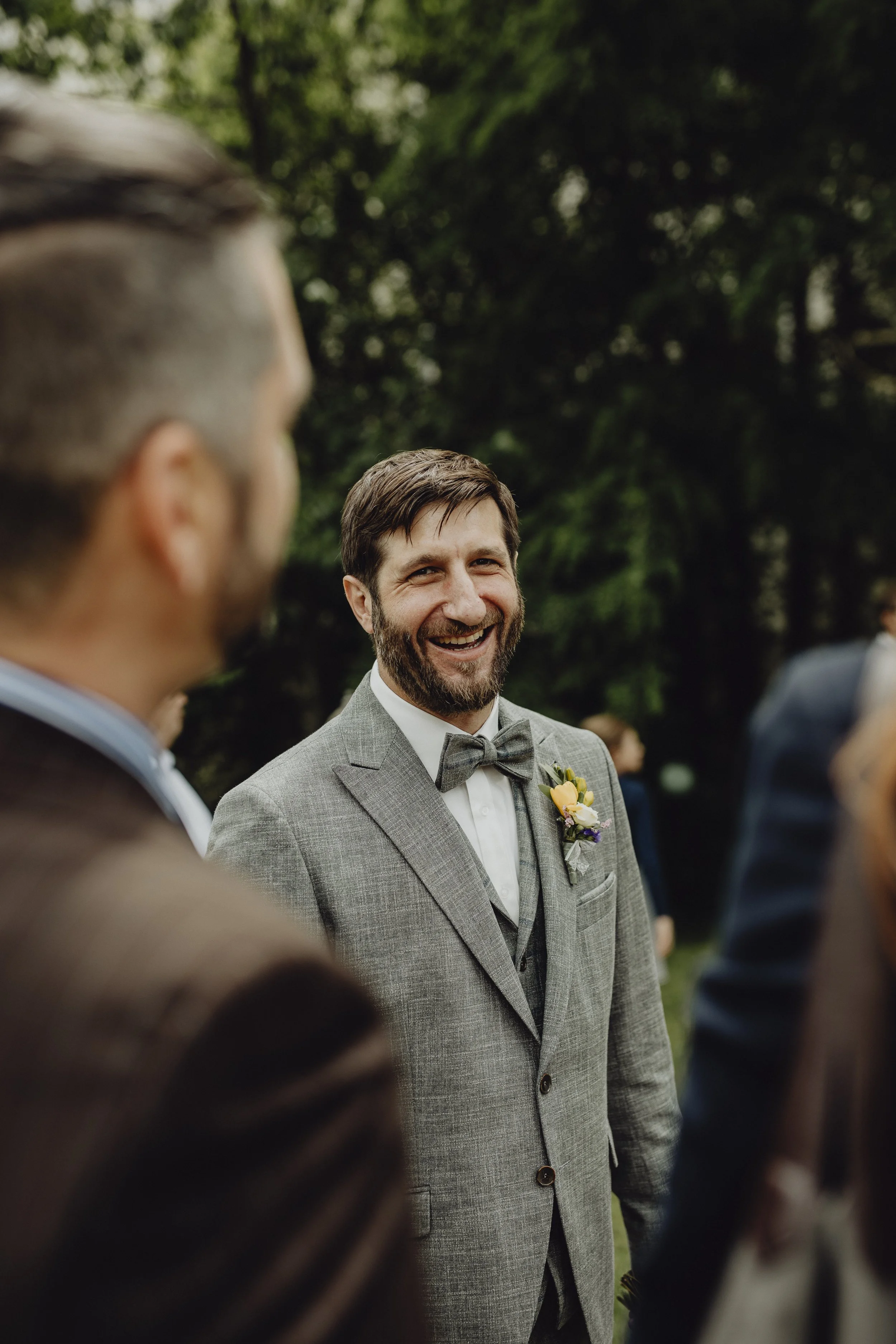 Ein lachender Bräutigam in einem Anzug mit Fliege, wahrscheinlich auf einer Hochzeitsfeier im Freien, umgeben von anderen Personen.