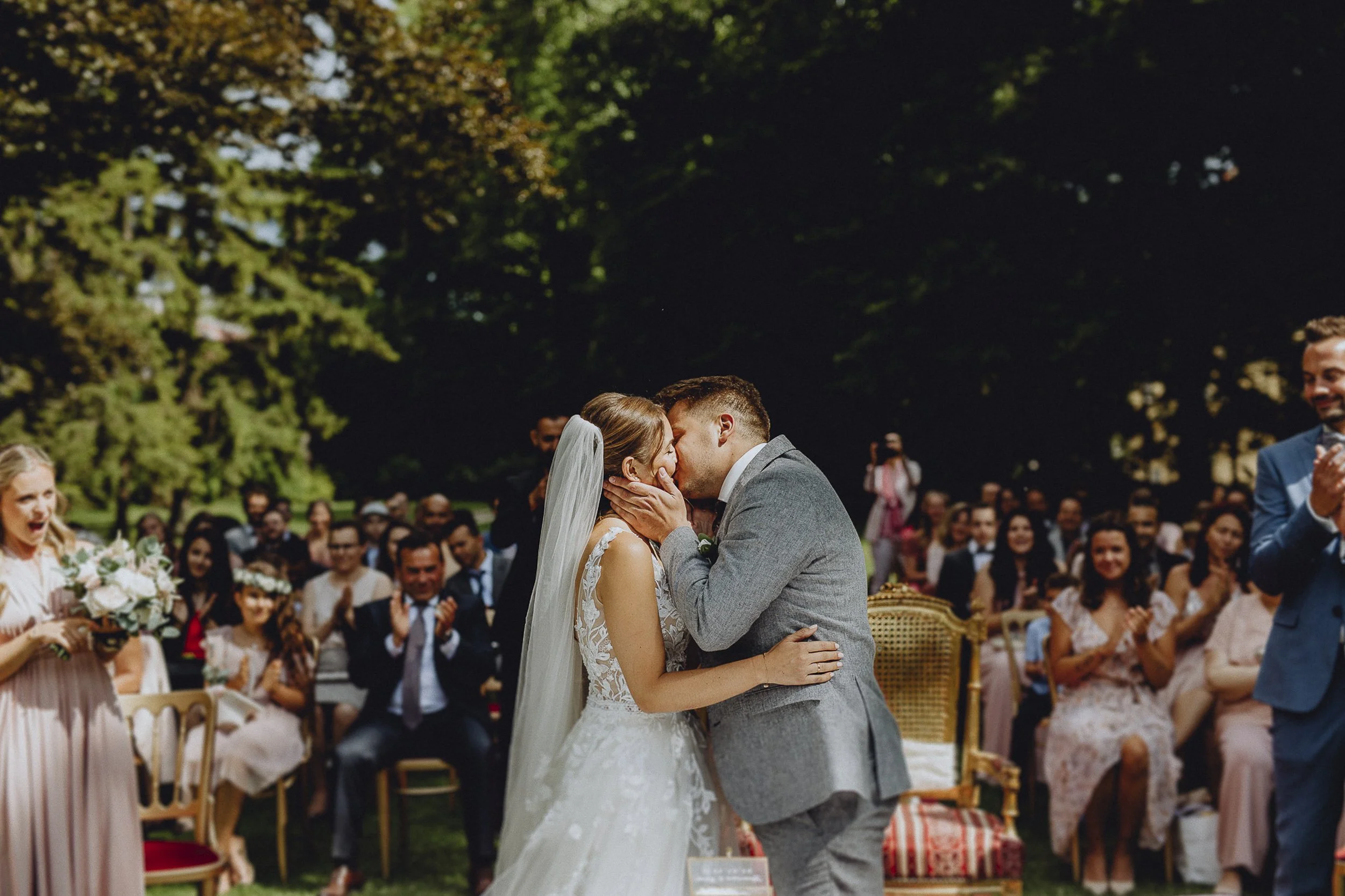 Ein Brautpaar bei der Hochzeit küsst sich, während sie sich umarmen, umgeben von lachenden Hochzeitsgästen im Freien.