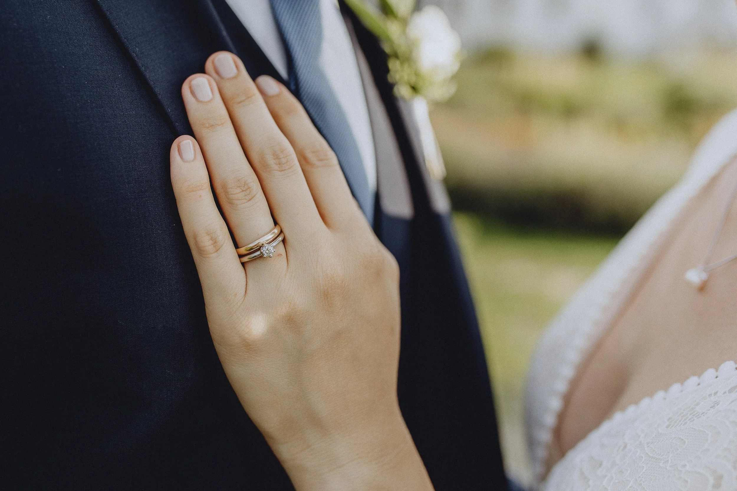 Nahaufnahme einer Hand mit Ehering, die sanft auf die Schulter eines Mannes mit dunklem Anzug und Krawatte gelegt wird, im Hintergrund unscharf.
