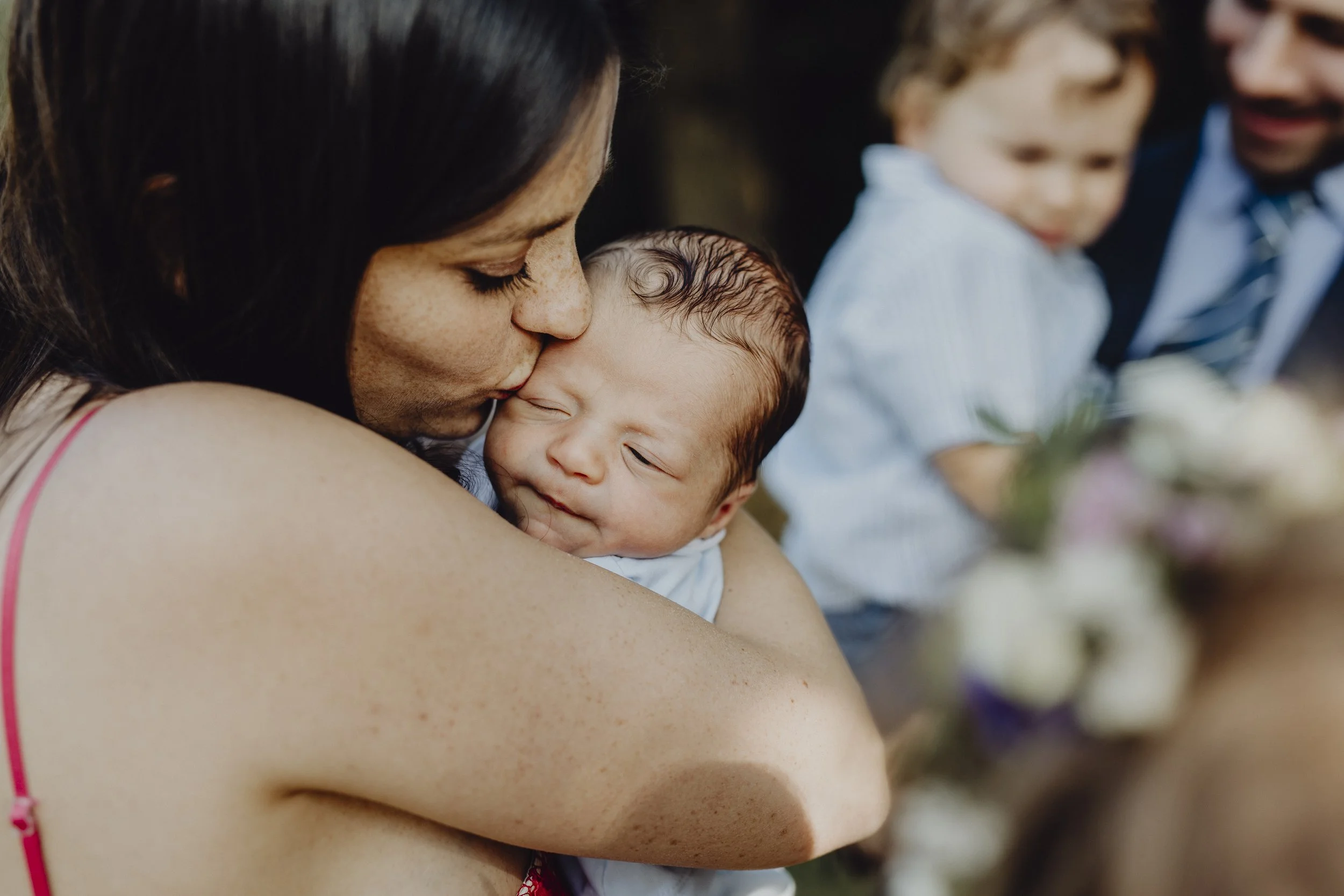 Frau hält schlafendes Baby nah an ihre Schulter, während sie es küsst, im Hintergrund lächelnder Mann und Kind bei einem Fest.