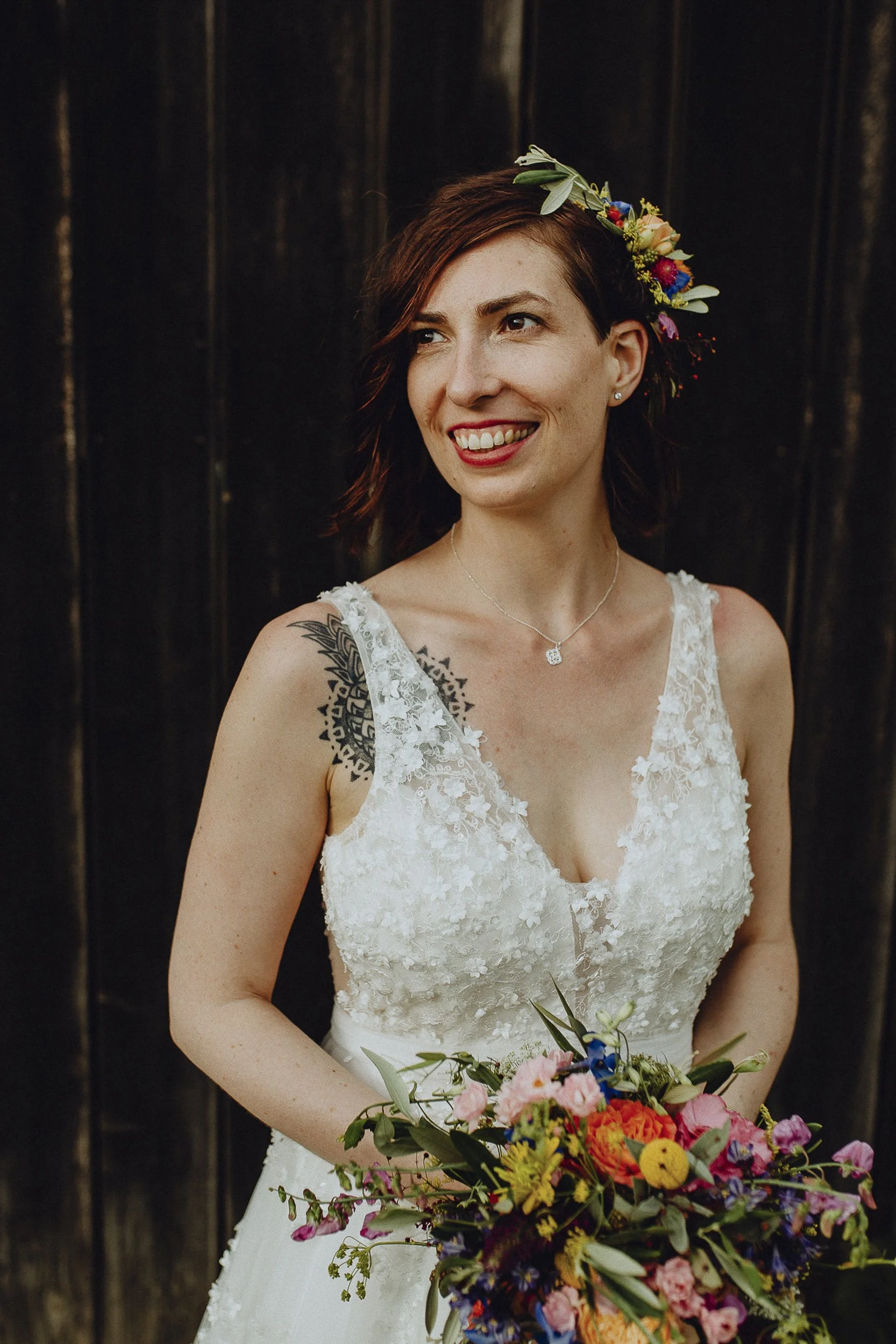 Eine lachende Frau in einem weißen Brautkleid mit Blumen im Haar, hält einen bunten Blumenstrauß, vor einem dunklen Holztor.