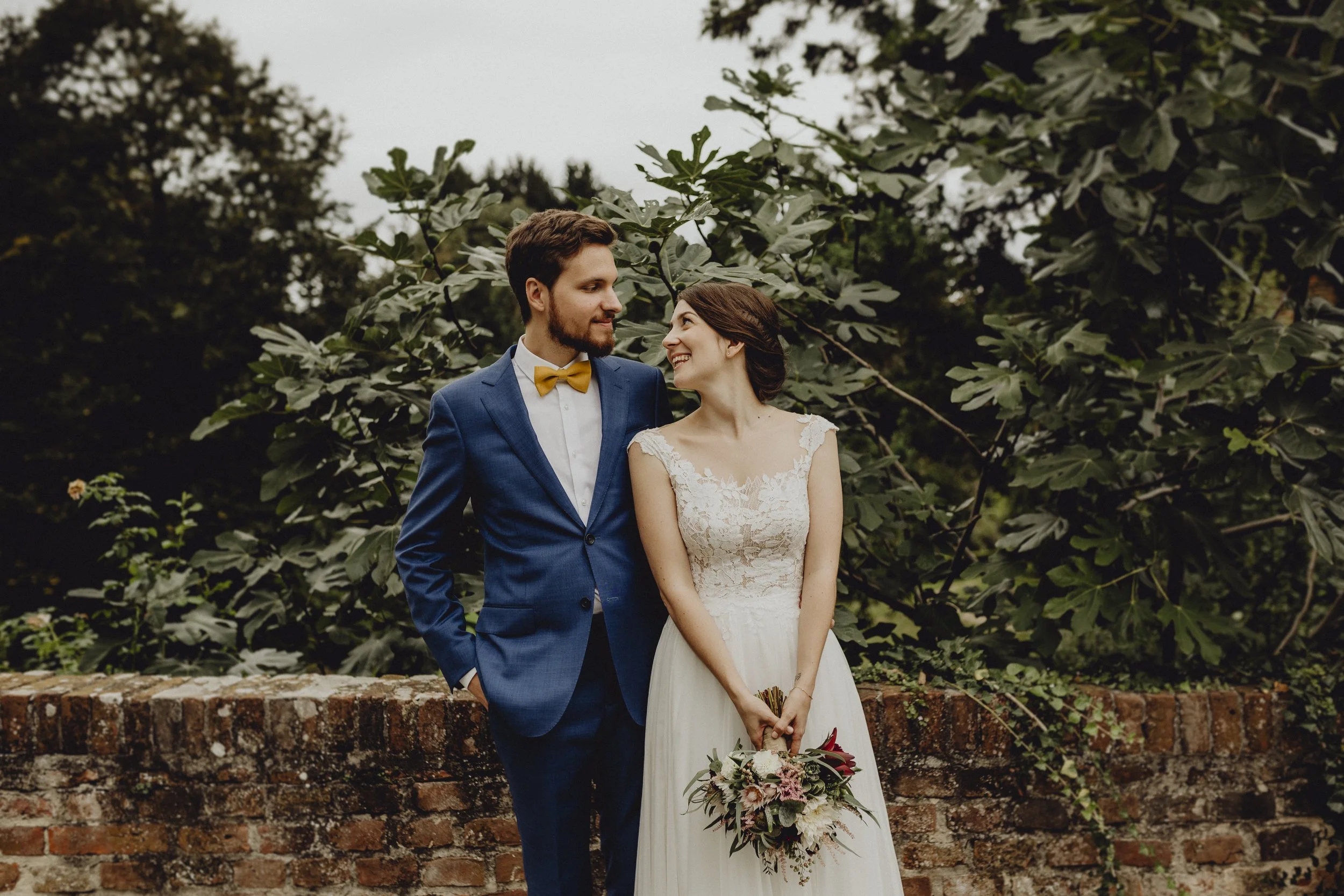 Ein Brautpaar steht vor einer alten Backsteinmauer, umgeben von grünen Büschen und Bäumen. Die Braut trägt ein weißes Spitzenkleid und hält einen Blumenstrauß. Der Bräutigam trägt einen blauen Anzug mit einer gelben Fliege.