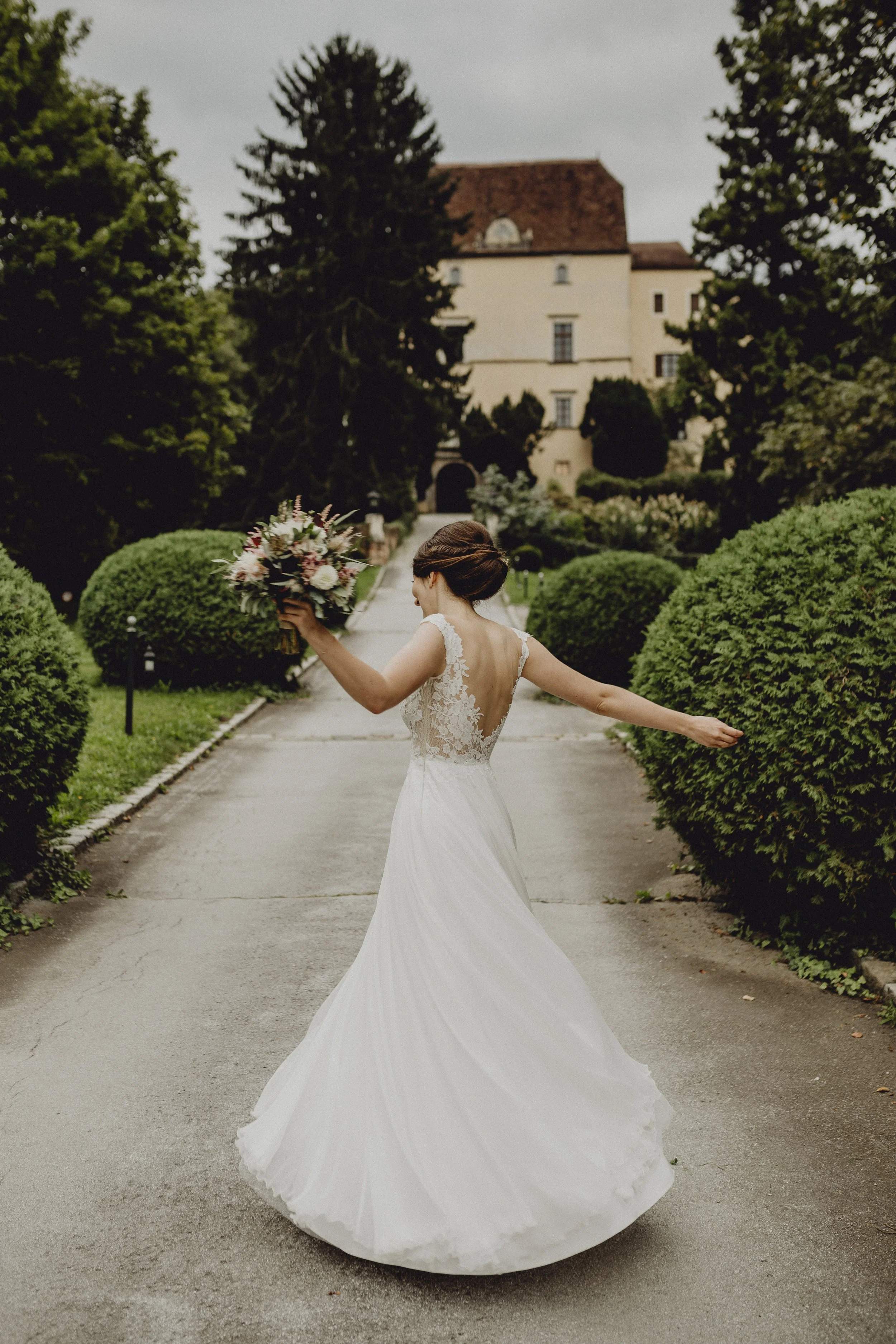 Braut in Hochzeitskleid läuft auf einem Weg in einem Garten, hält Blumenstrauß in einer Hand, und hat die andere Hand ausgebreitet, im Hintergrund ein altes Gebäude und Bäume.