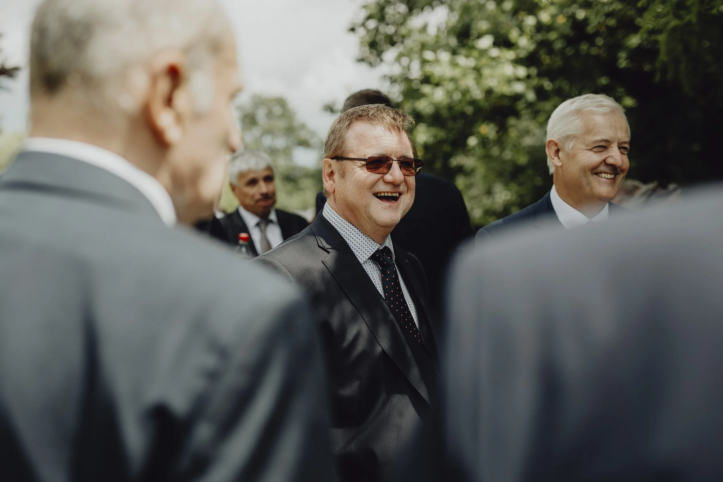 Gruppe von Männern im Anzug beim Außenmeeting, lachend und im Gespräch, mit grüner Natur im Hintergrund.
