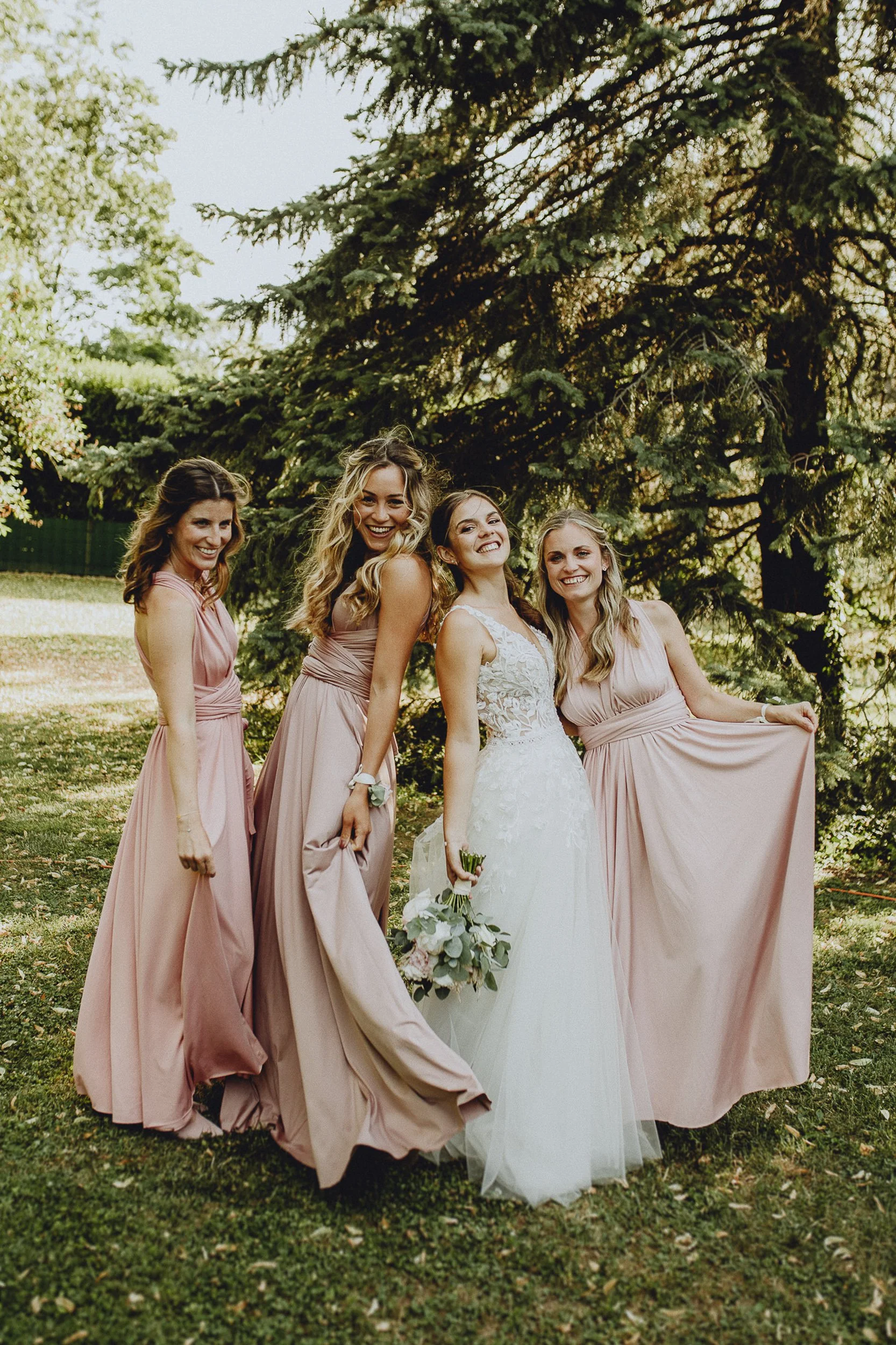 Vier Frauen, eine in einem weißen Brautkleid, die anderen in rosa Brautjungfernkleidern, lachen und posieren im Freien vor einem großen Baum auf einer grünen Wiese an einem sonnigen Tag.