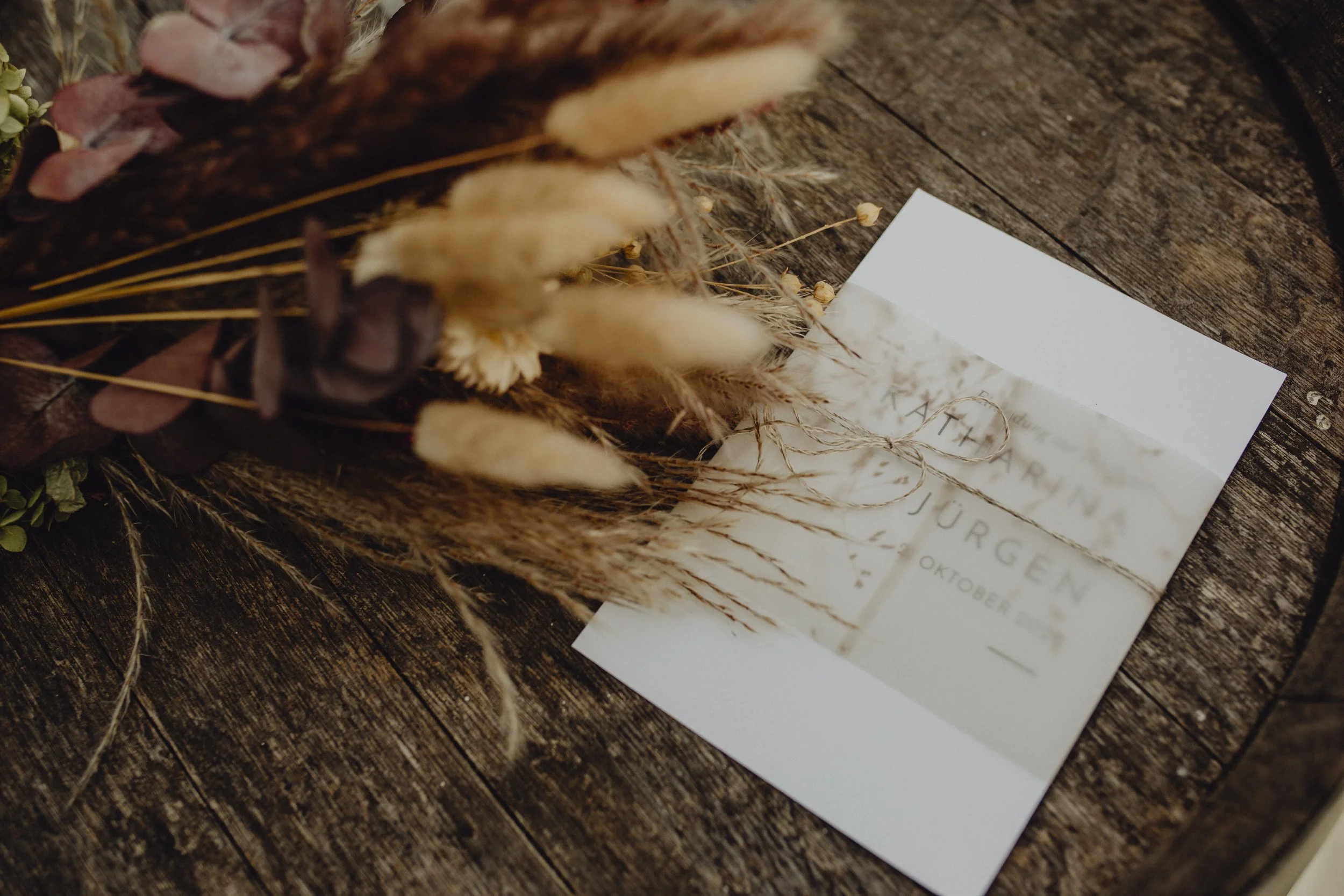 Eine Hochzeitskarte auf einem rustikalen Holzoberteil, umwickelt mit einem Juteschnur, neben einer Blumen- und Getreide-Arrangements.