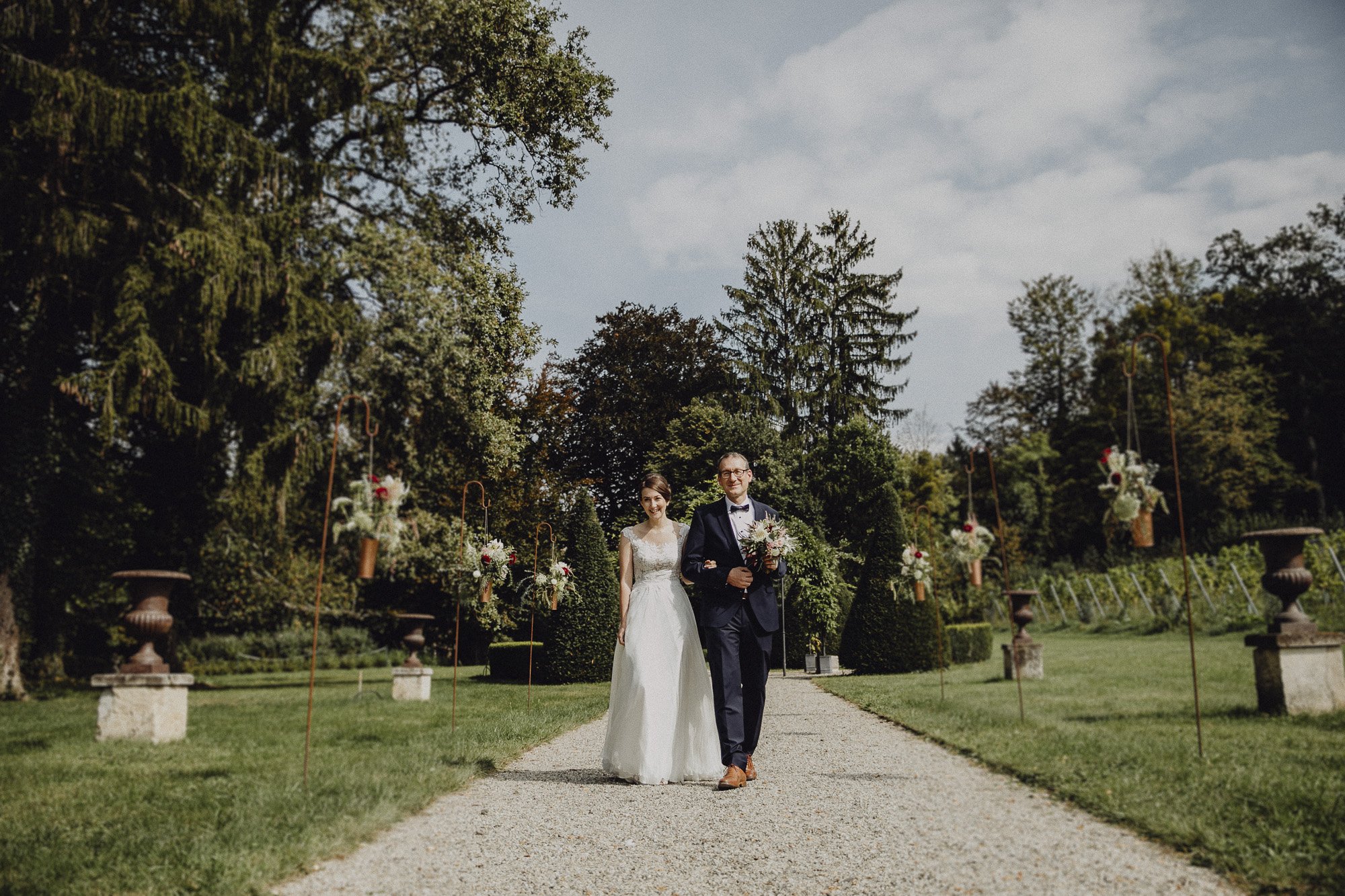 Braut in weißem Kleid und Bräutigam in dunklem Anzug gehen an einer Gartenstraße entlang, mit Blumen und Pflanzen im Hintergrund.