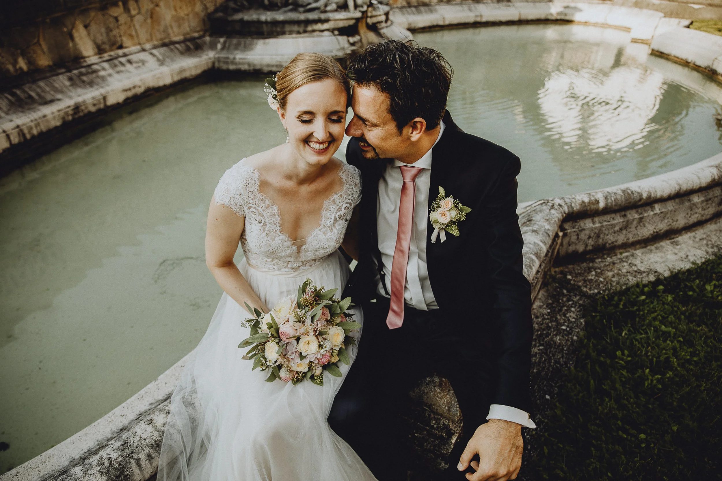 Ein glückliches Brautpaar sitzt an einem Brunnen. Die Braut trägt ein weißes Kleid mit Spitze und hält einen Blumenstrauß. Beide lachen, während er ihr etwas ins Ohr flüstert.