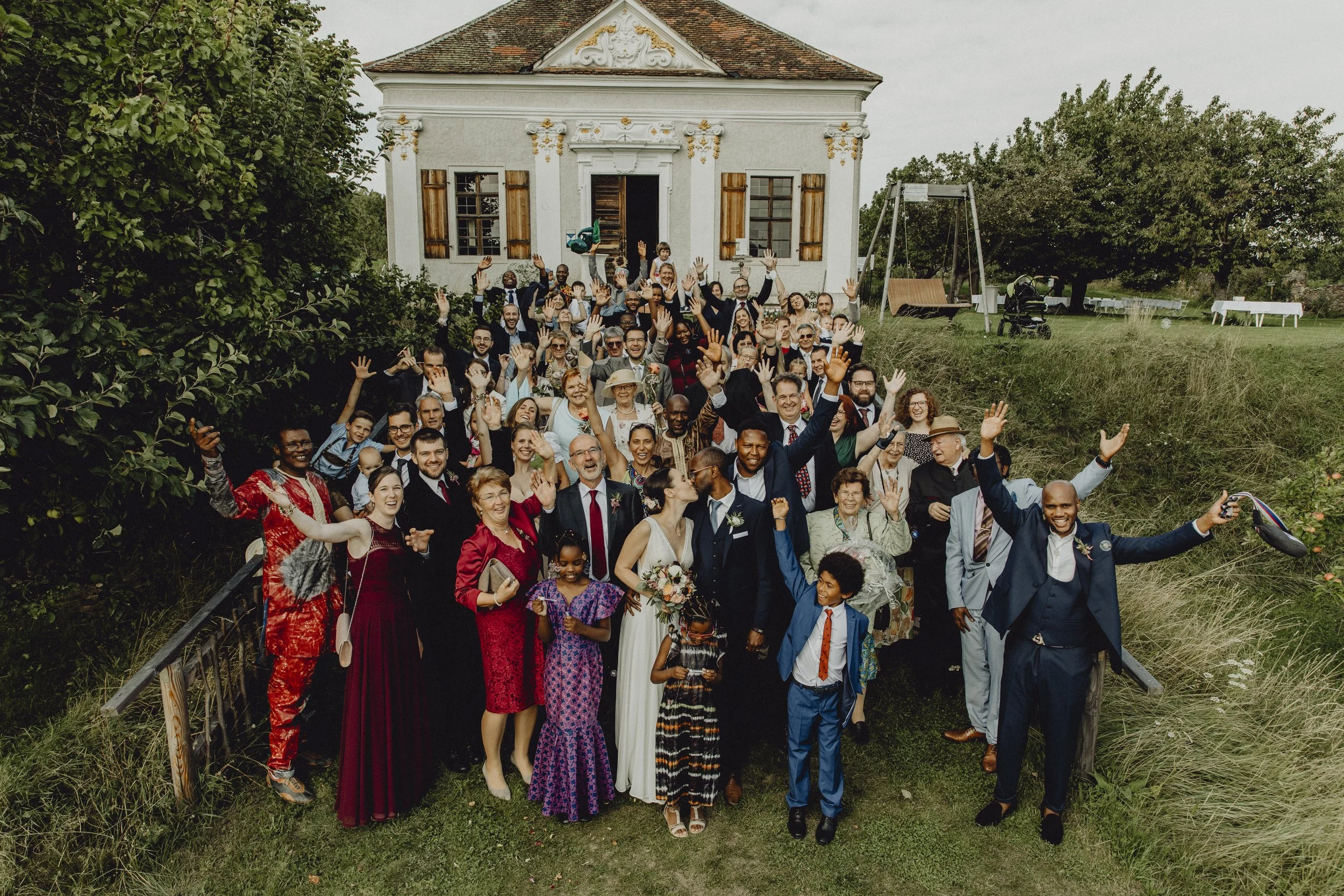 Gruppenfoto einer Hochzeit auf einer Rasenfläche vor einem weißen Gebäude, einige winken, andere posieren für das Foto, im Hintergrund Bäume und viel grün.